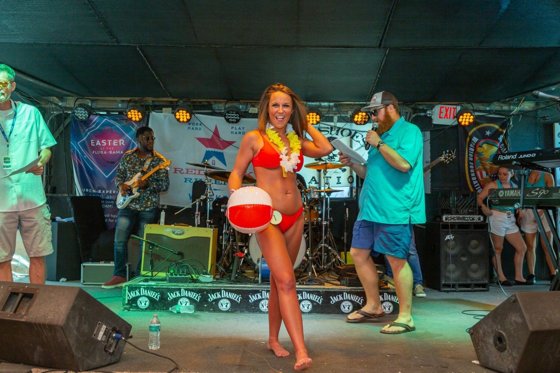 A person in a red bikini holds a beach ball on a stage with a band, while an announcer stands next to them with a mic.