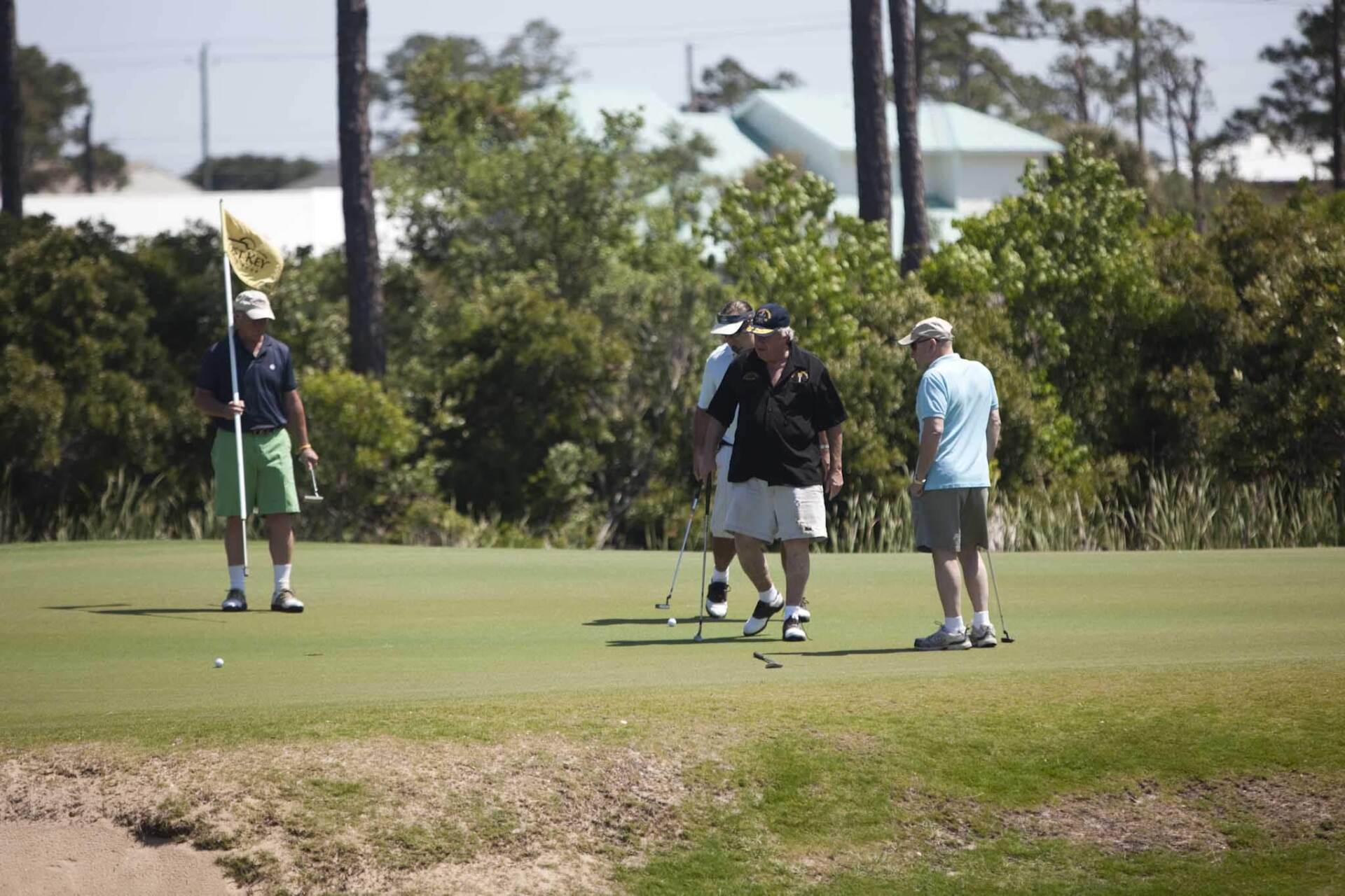Four people on a sunny golf green; one person holds a flag while three others walk and talk with golf clubs.