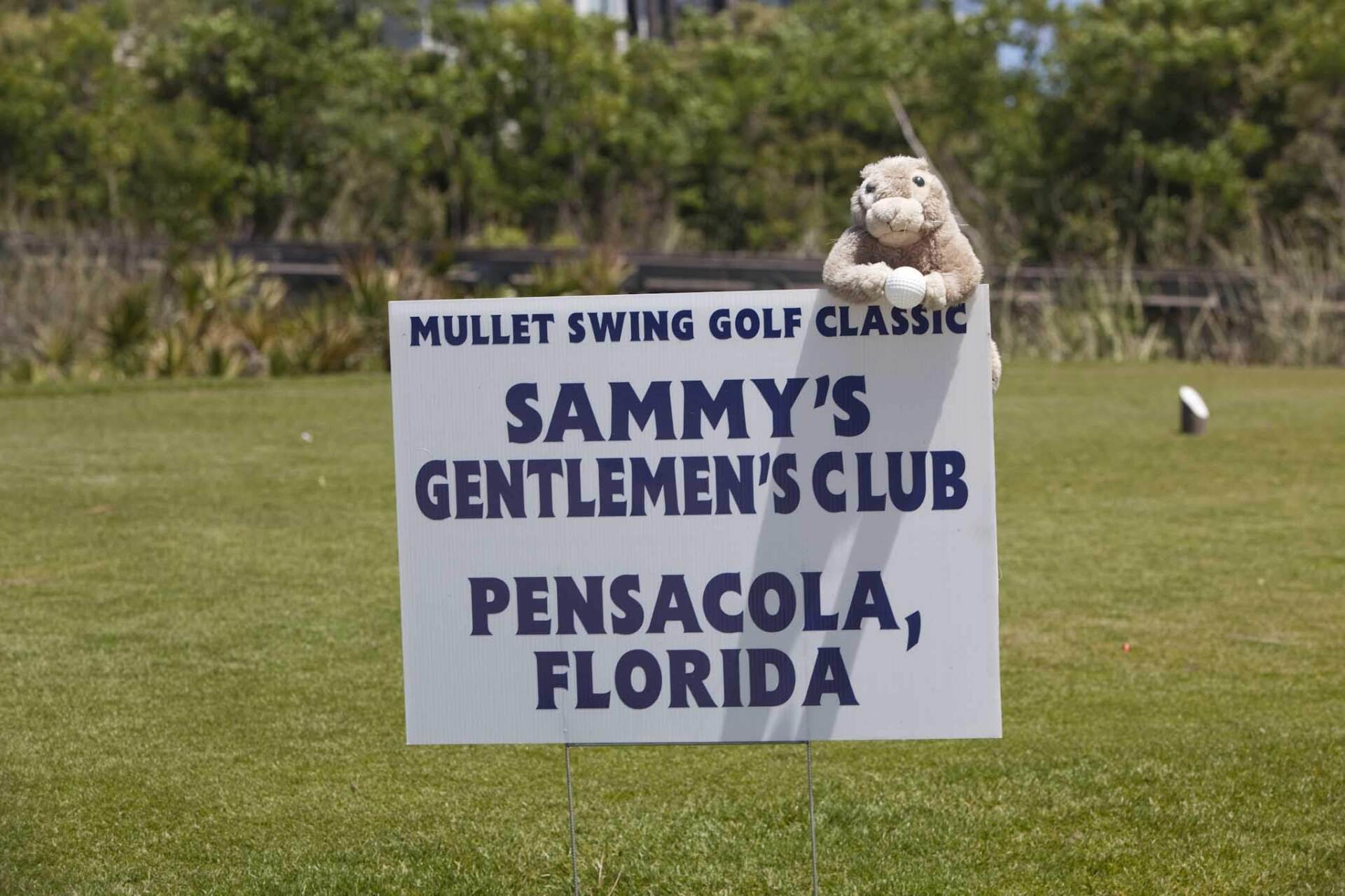 A sign for the Mullet Swing Golf Classic in Pensacola, Florida, featuring Sammy’s Gentlemen’s Club, held on a golf course.
