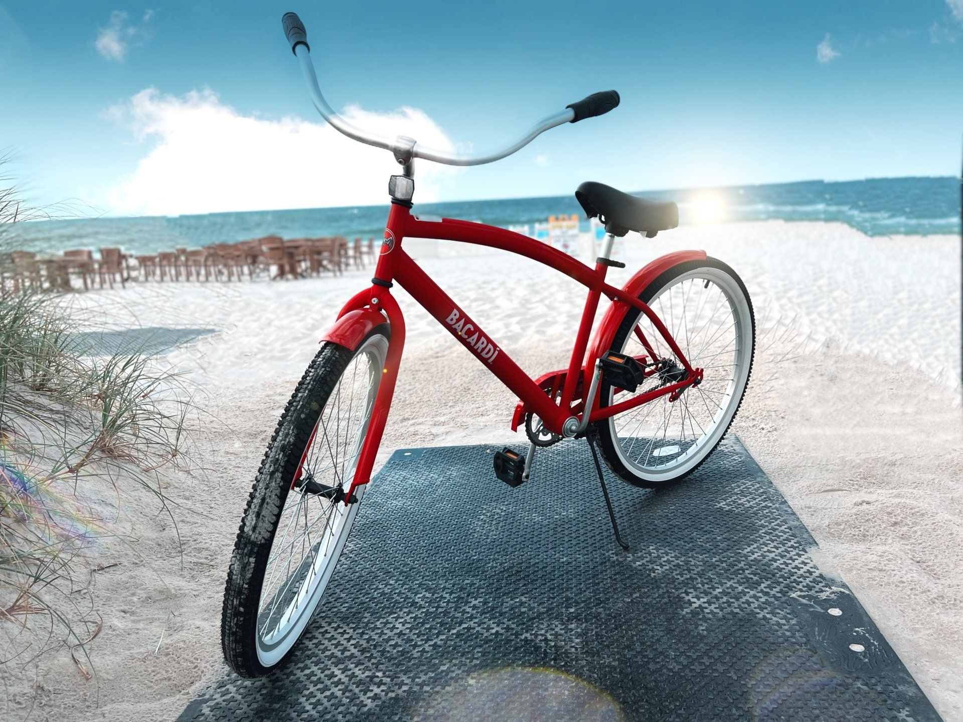 A bright red cruiser-style bicycle stands on a textured black mat on a white sandy beach with the ocean in the background.