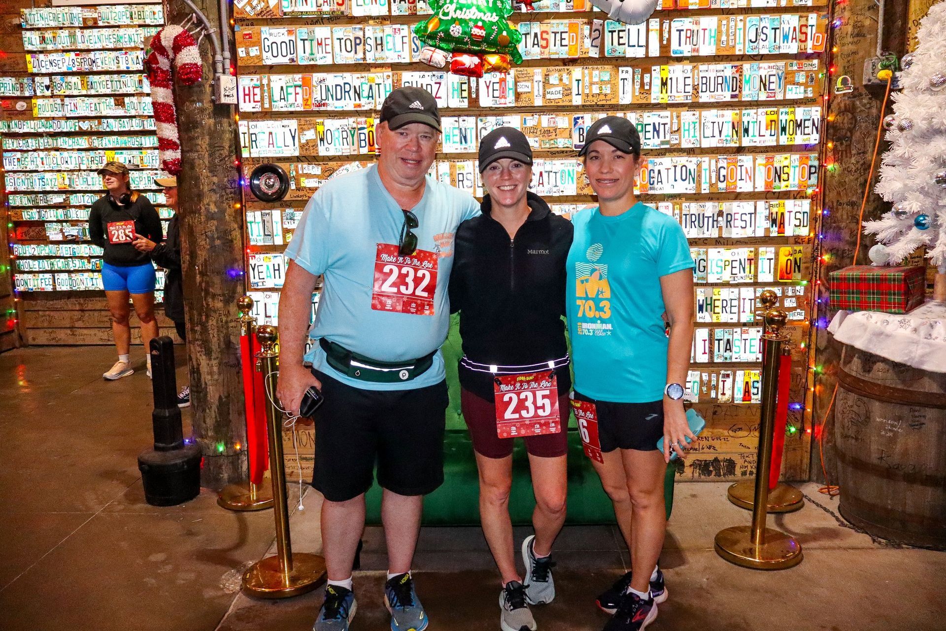 Three people pose in front of a wall of license plates, wearing race bibs 232, 235, and 233, respectively.