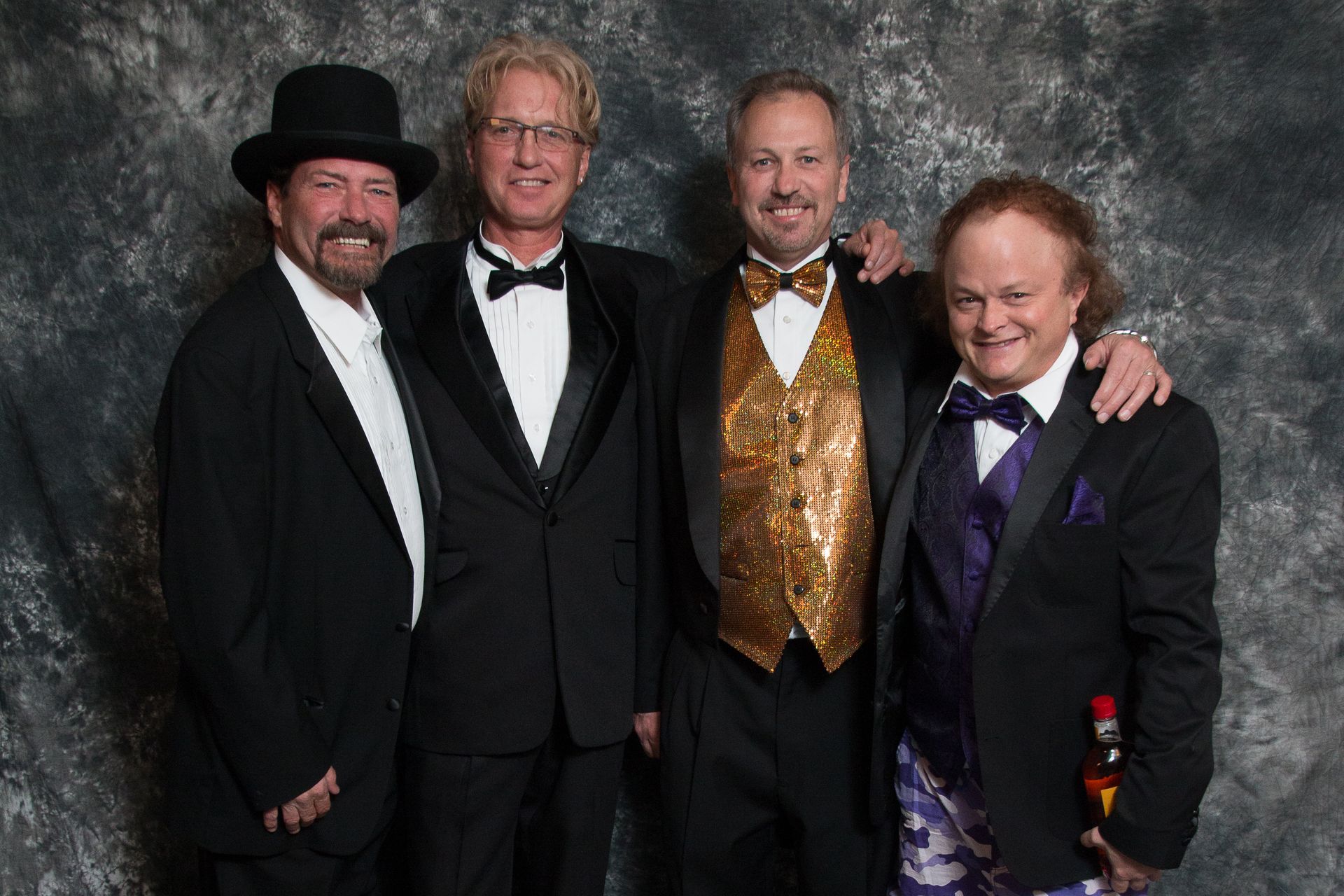 Four men in formal attire, including tuxedos and a top hat, pose together in front of a gray mottled backdrop.