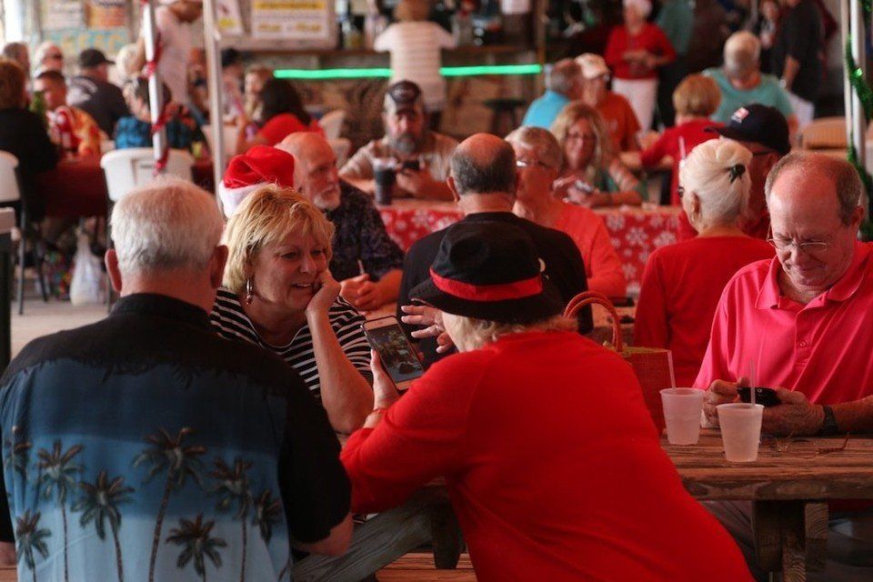A group of people sit at wooden tables in a festive, crowded social setting, some eating or looking at their phones.