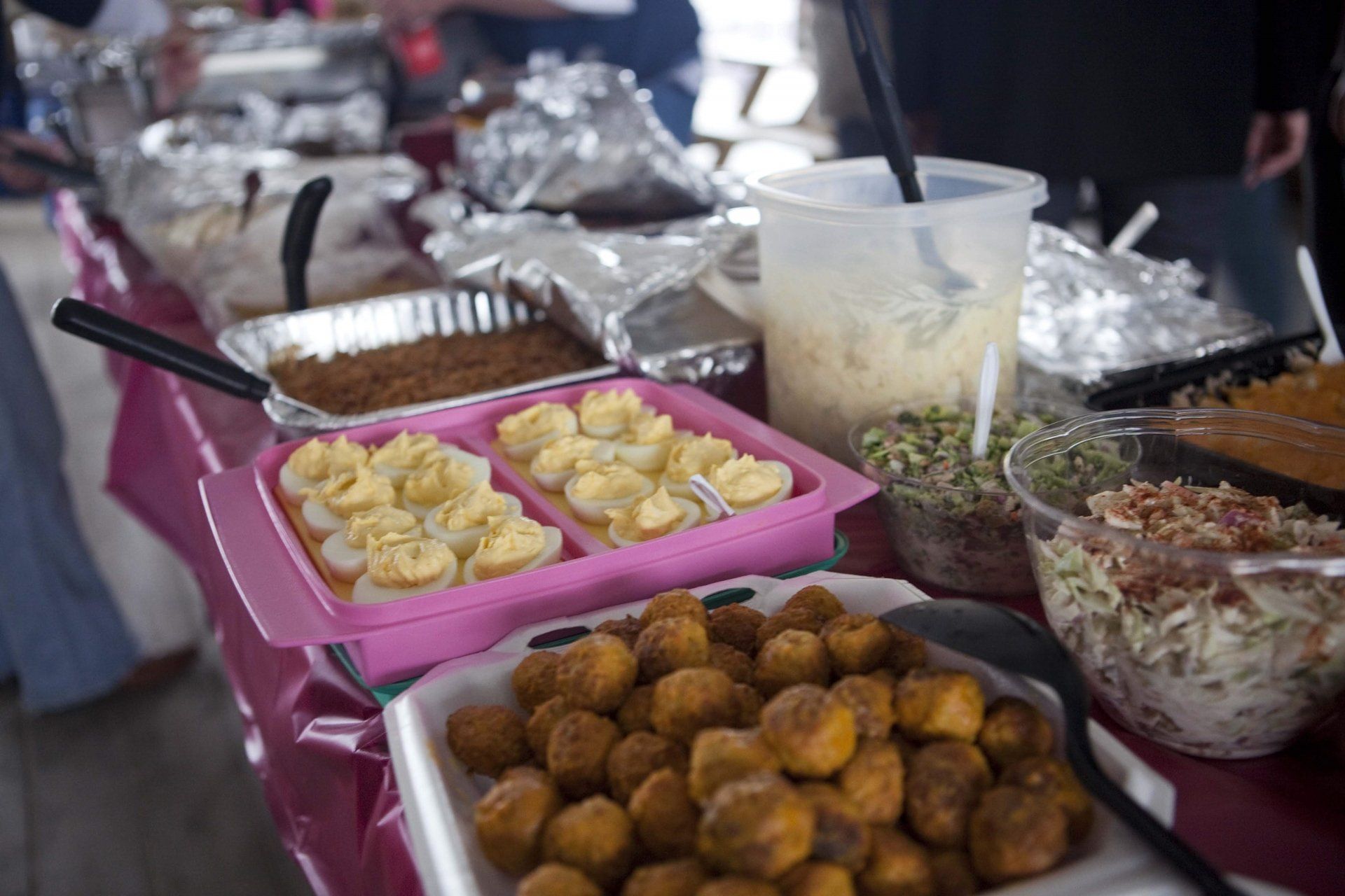 A food buffet with deviled eggs, round fried items, salads, and foil-covered dishes on a pink tablecloth.