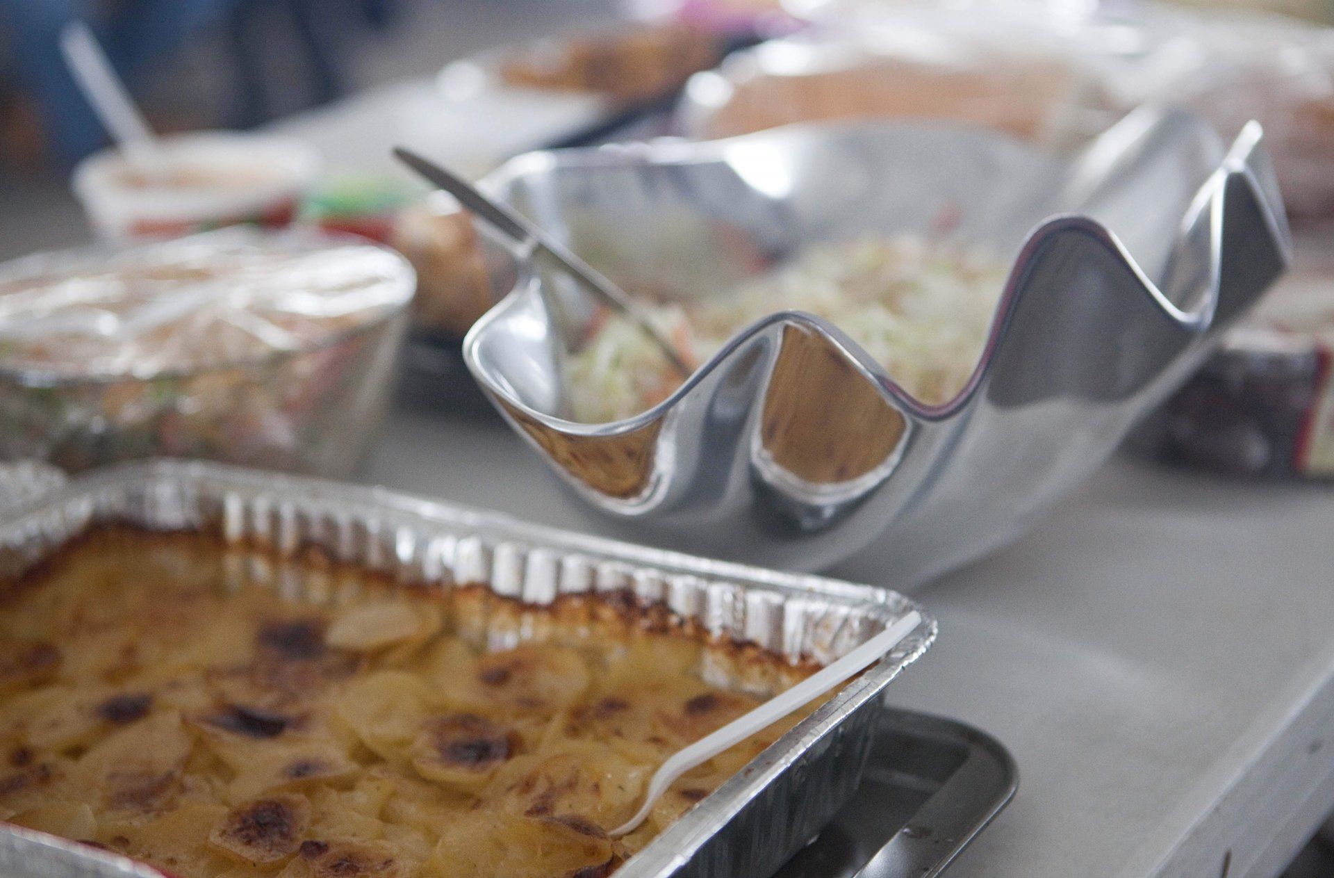 A table with a metal bowl of coleslaw and a foil tray of scalloped potatoes, with other dishes blurred in the background.