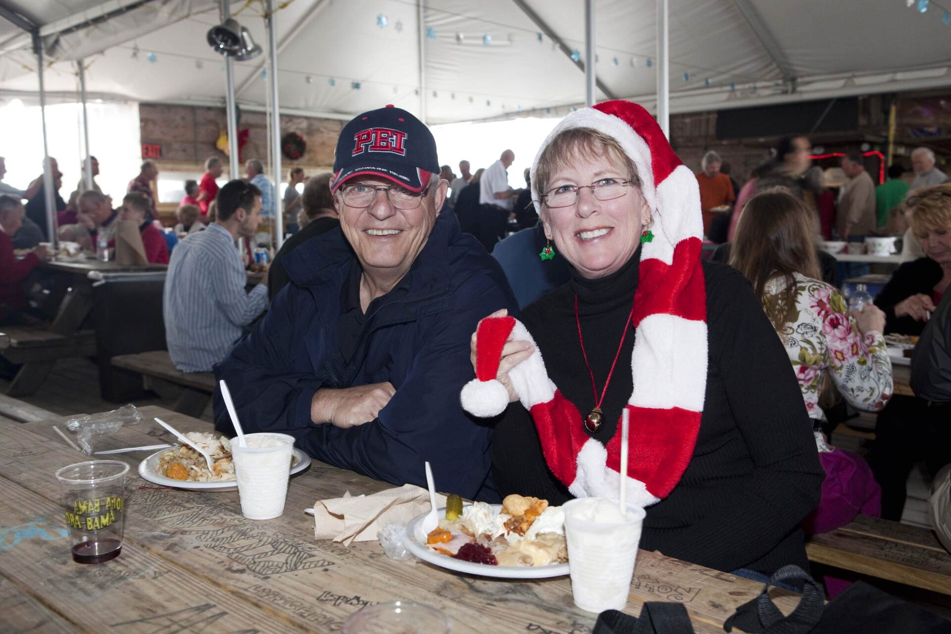 A smiling couple sits at a rustic table in a large tented event space, enjoying a meal and wearing festive holiday gear.