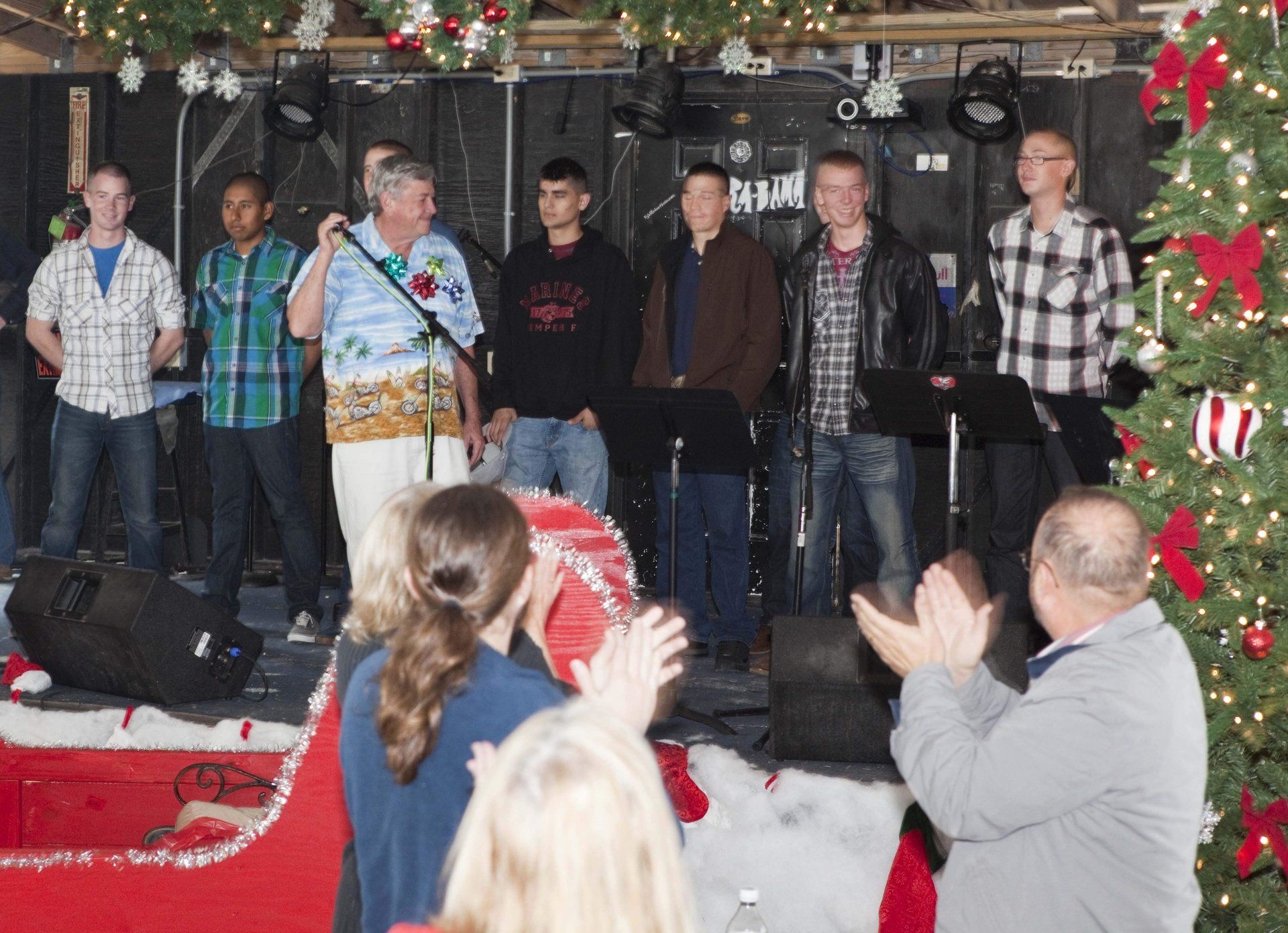 A group stands on a stage decorated with holiday lights and a tree, while audience members clap in the foreground.
