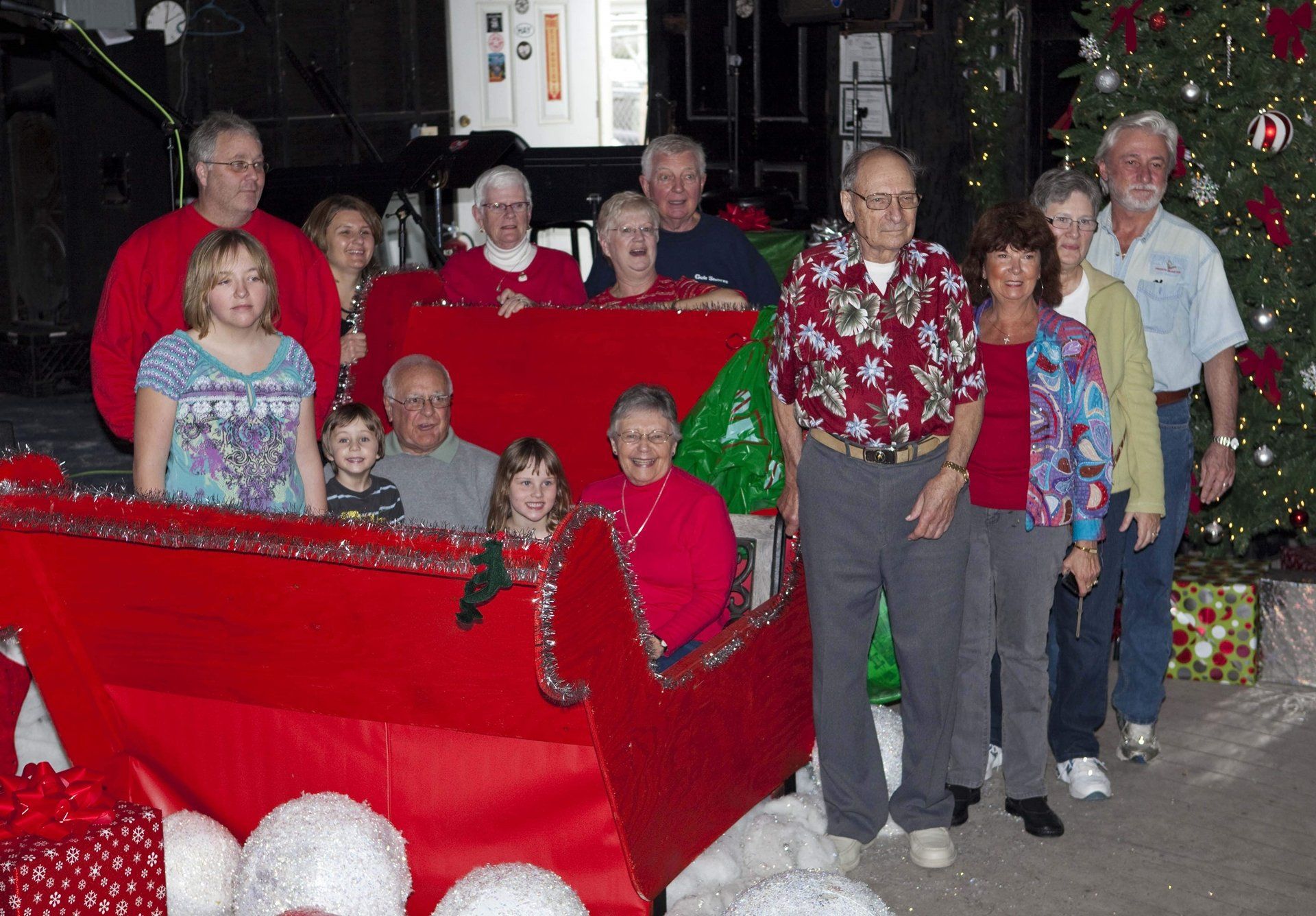 A group of people gathers around a large red sleigh in a holiday-themed setting with a decorated Christmas tree.