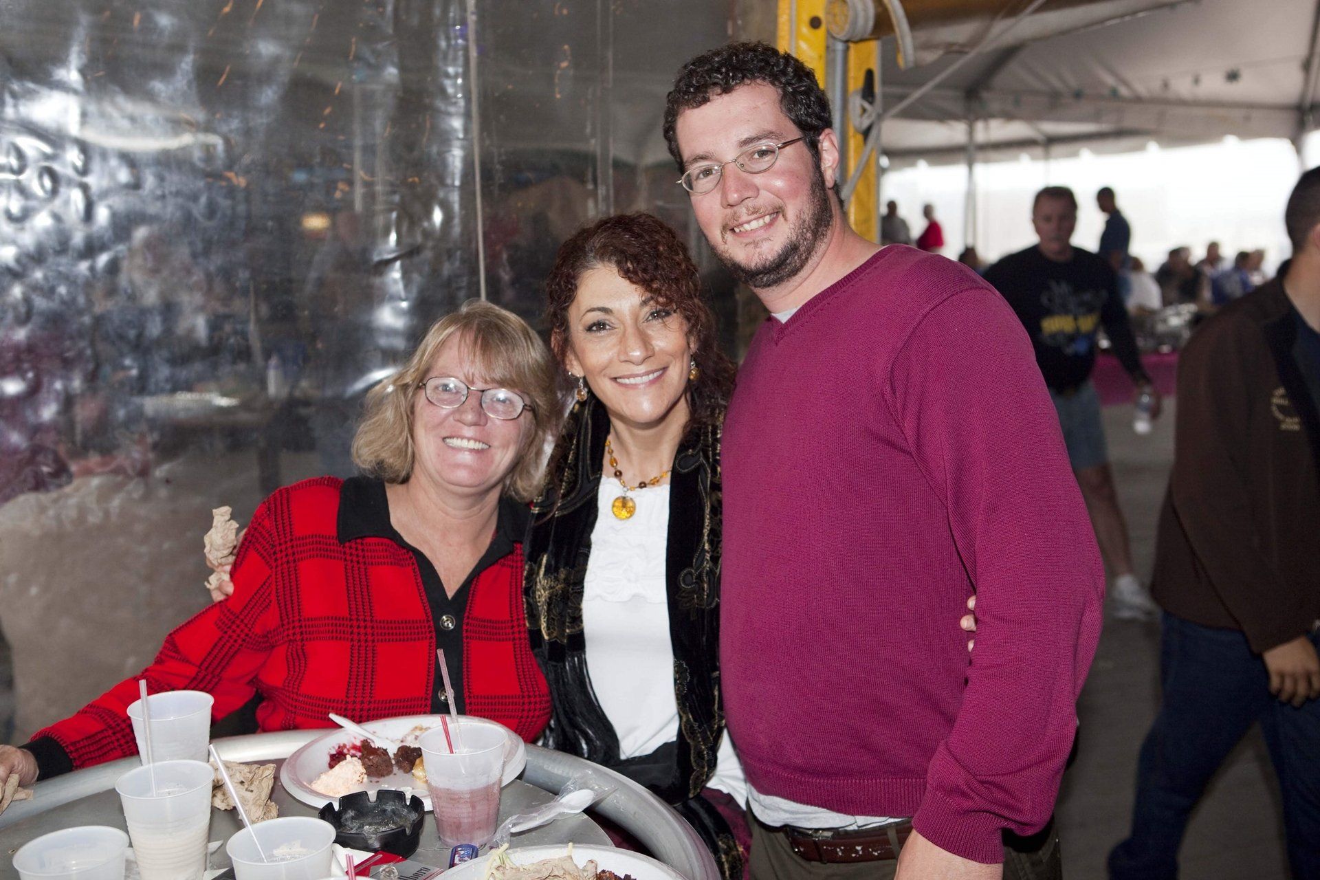 Three people smile together at a table in a tented, outdoor event space.