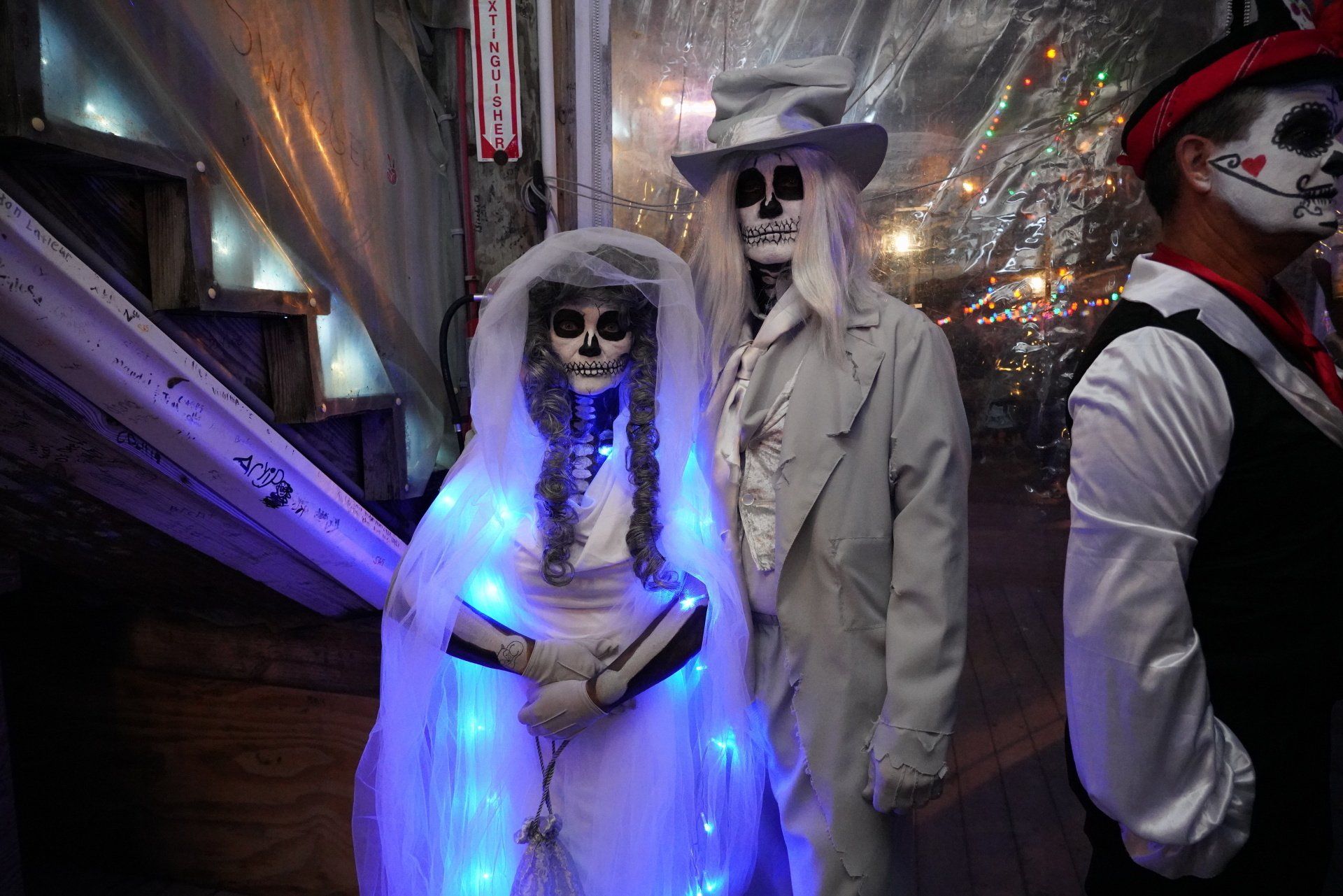 Three people in skeletal makeup and formal attire, including a glowing white veil, standing in a dimly lit, festive room.
