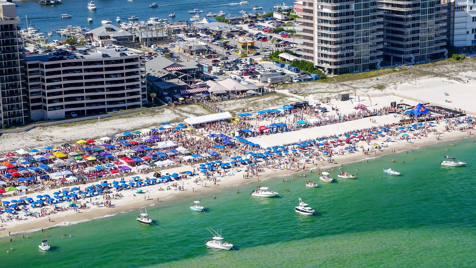 An aerial view of a crowded beach filled with colorful umbrellas and tents, with several boats anchored in the water.
