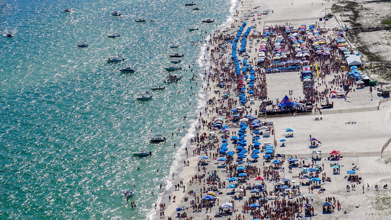 Aerial view of a sunny, crowded beach with blue umbrellas, numerous boats offshore, and people enjoying the water and sand.