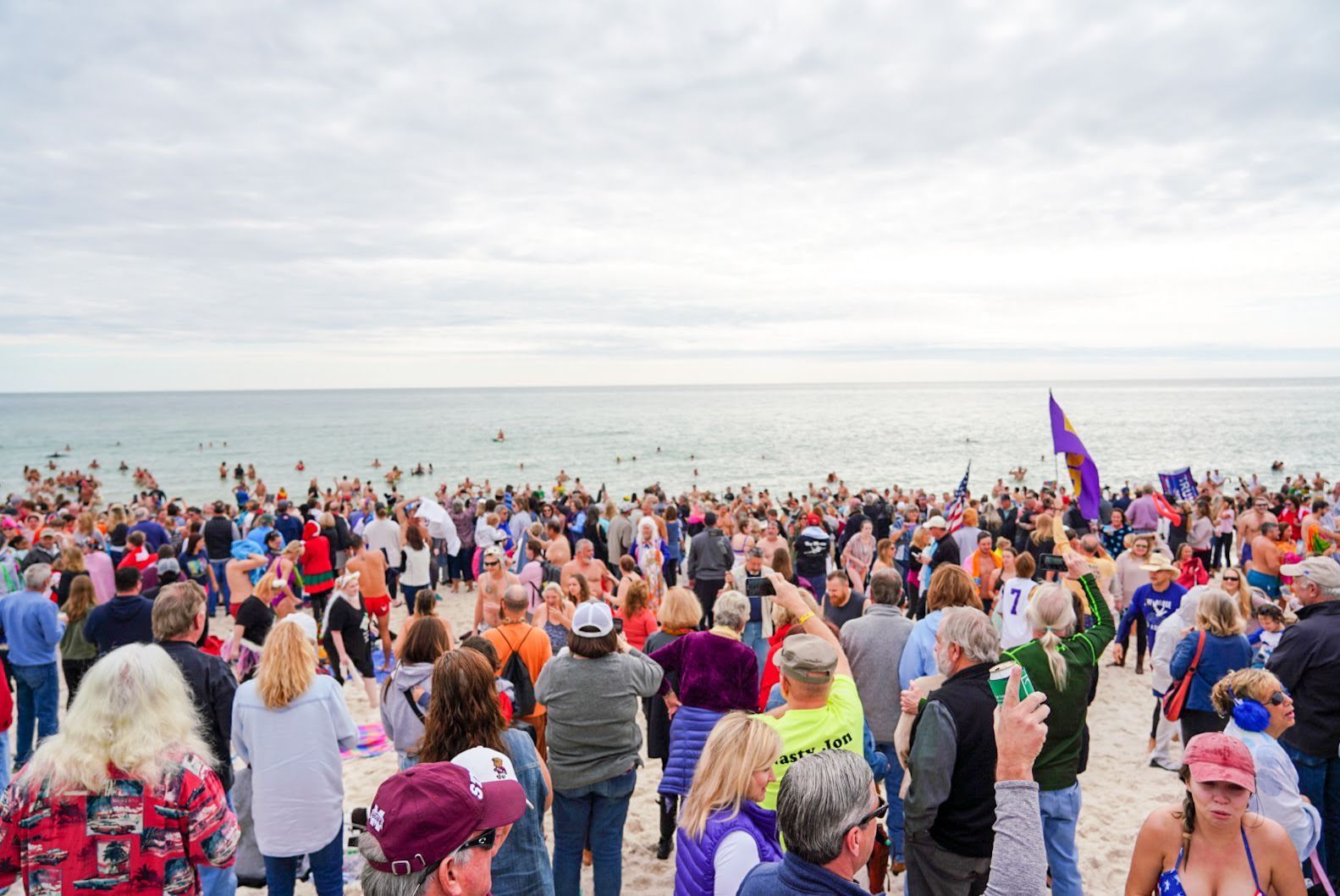 A large crowd gathers on a sandy beach, with many people preparing to enter the ocean for a winter swim under a cloudy sky.