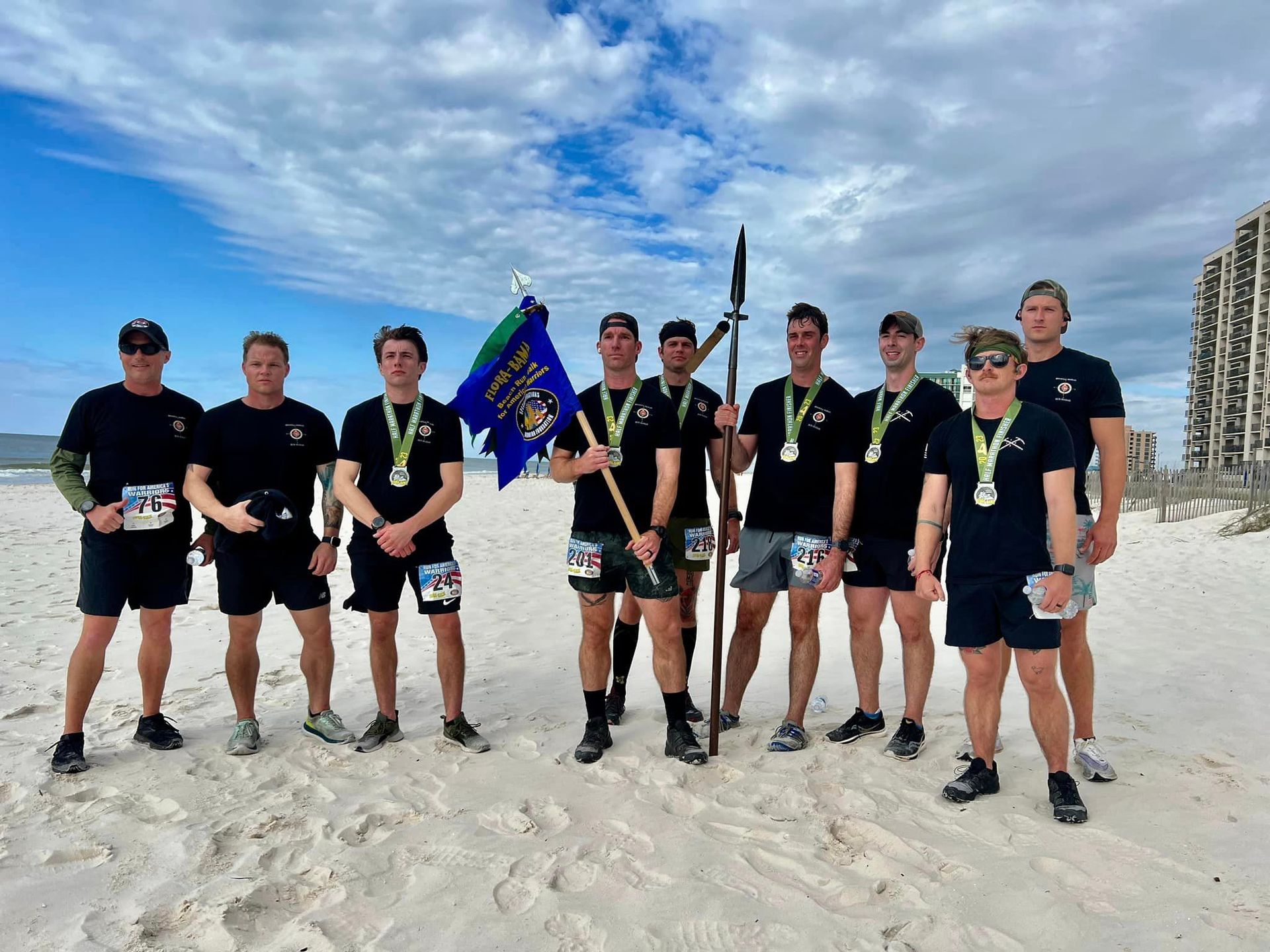 Ten people in matching dark athletic gear stand on a sandy beach, some holding medals, a flag, and a tall pole.