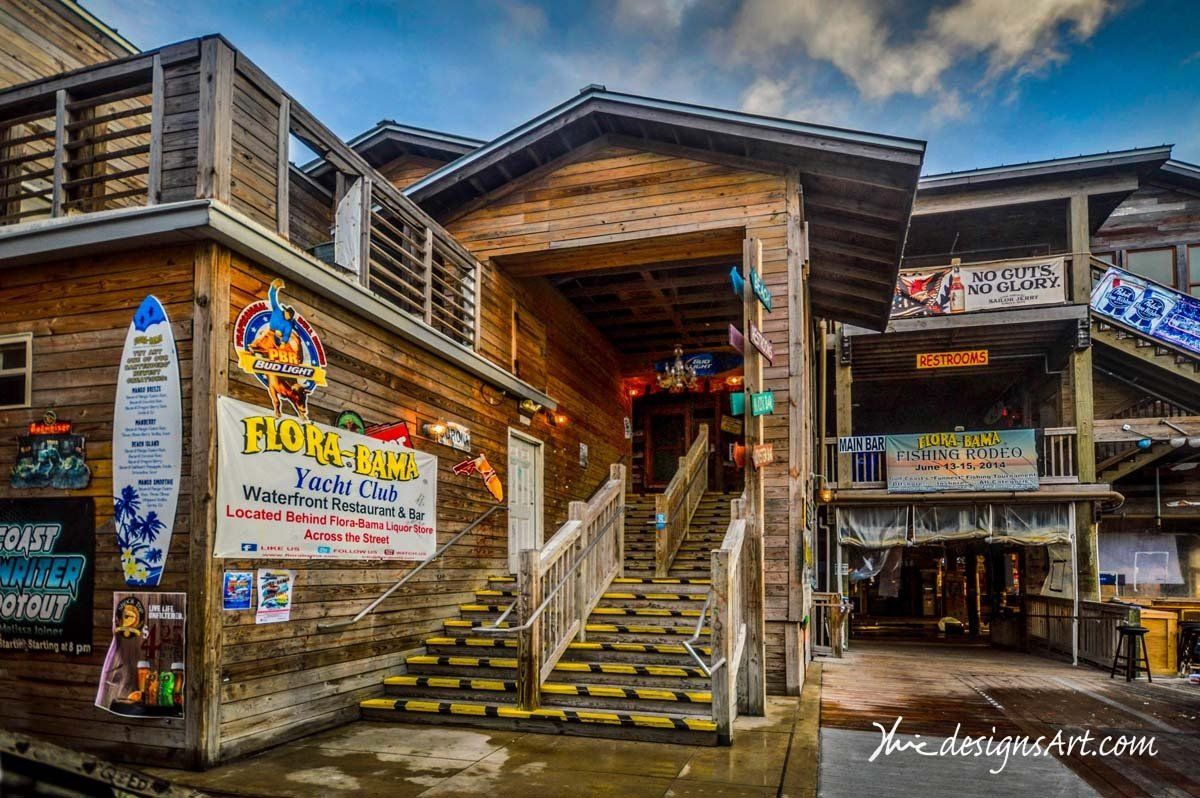 A wooden exterior of the Flora-Bama lounge with stairs leading up to an entrance decorated with a surfboard and signs.