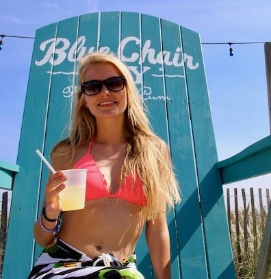 A person in a pink bikini top and patterned wrap smiles while holding a drink in a large, teal