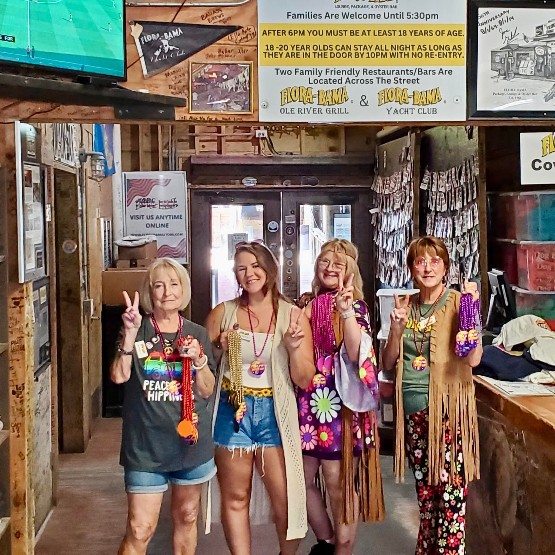 Four people in colorful, bohemian-style clothing pose with peace signs inside a rustic tavern.