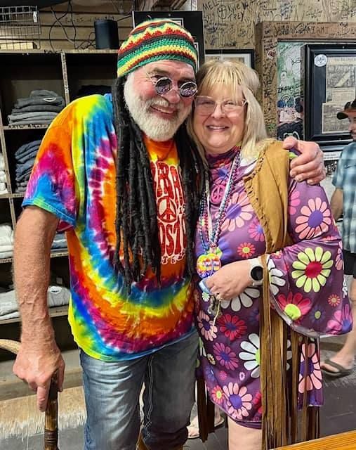 A smiling couple in 1960s-style tie-dye and floral outfits poses together inside a store with wall decorations.