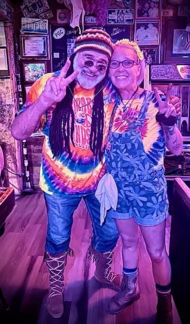 Two people in tie-dye shirts pose with peace signs in a dimly lit, decor-filled room; one wears dreadlocks and a beanie.