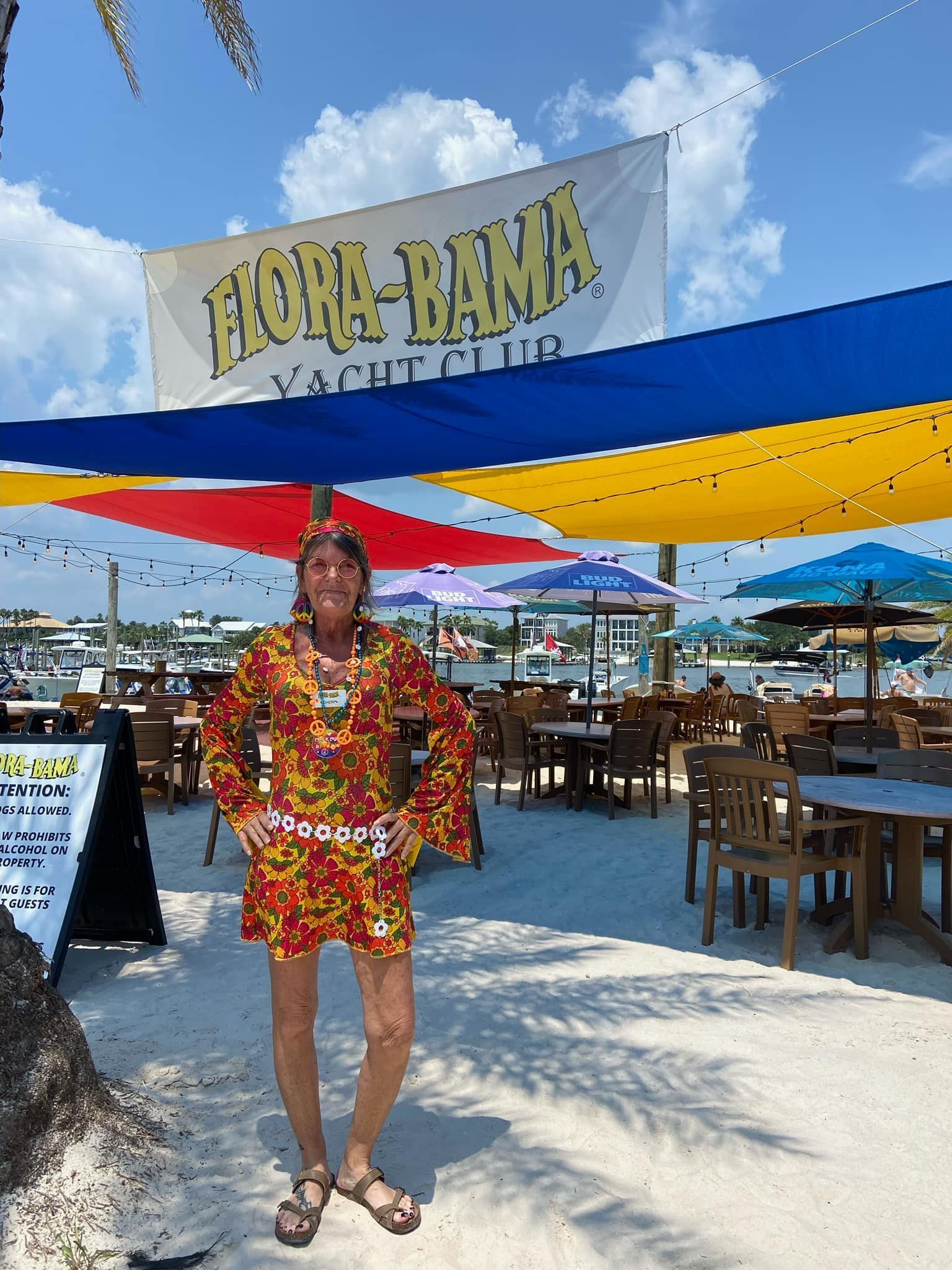A person in a bright floral outfit stands on a sandy patio beneath colorful sun shades at Flora-Bama Yacht Club.
