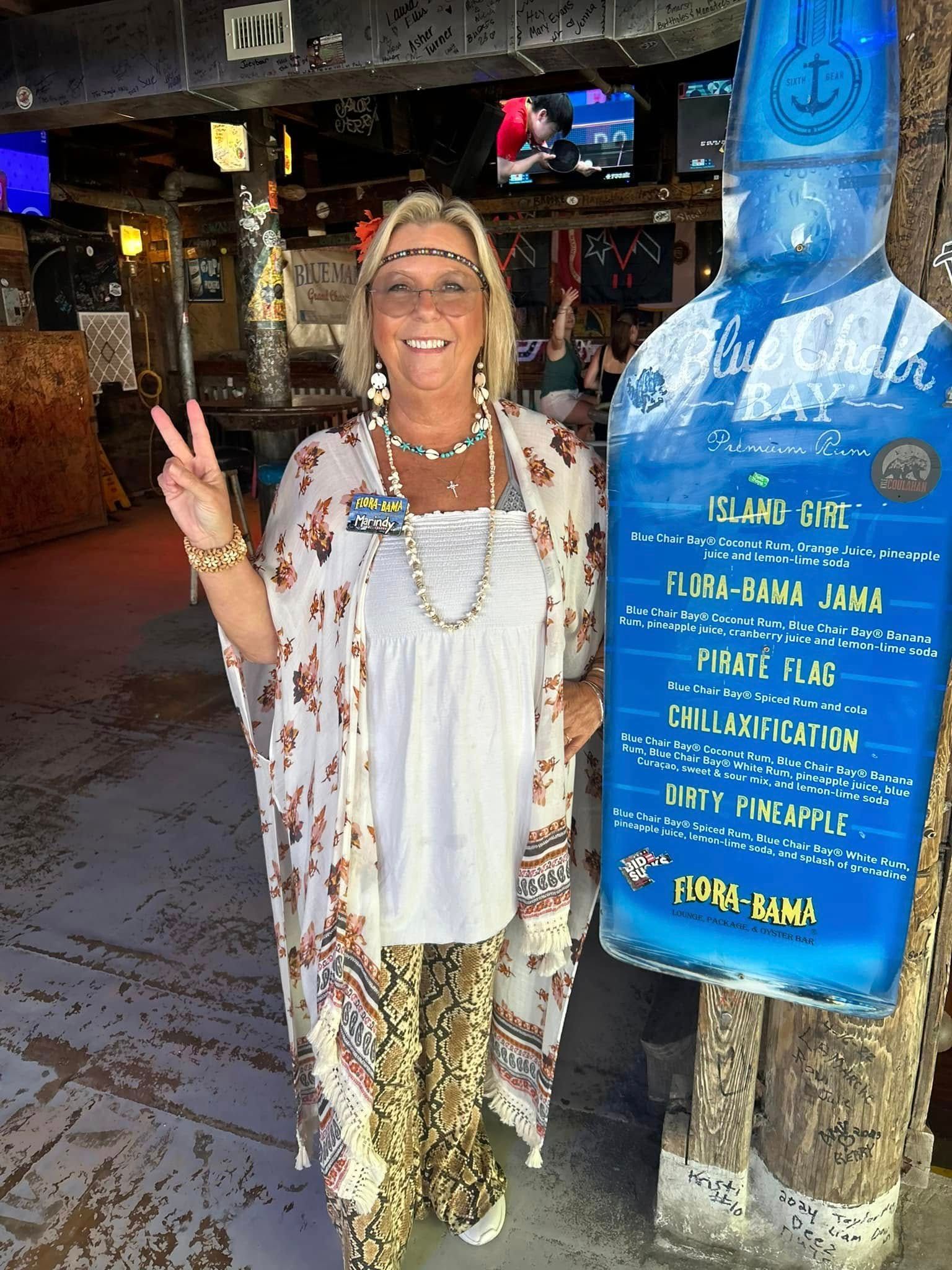 A smiling woman in a patterned kimono and headband makes a peace sign next to a blue menu board in a restaurant.