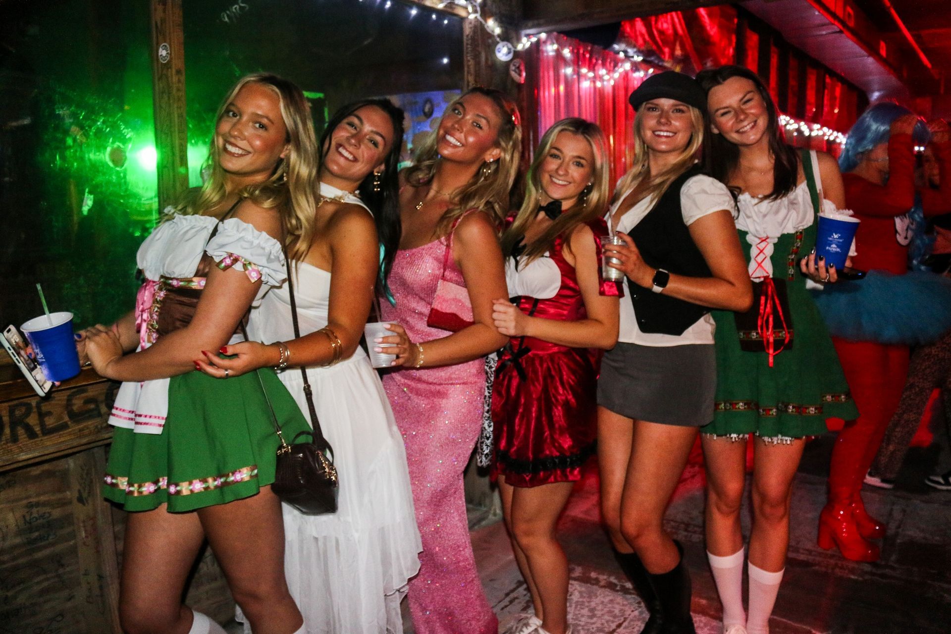 Six people pose together at a bar, some wearing costumes, including green skirts and a pink dress, in a dimly lit setting.