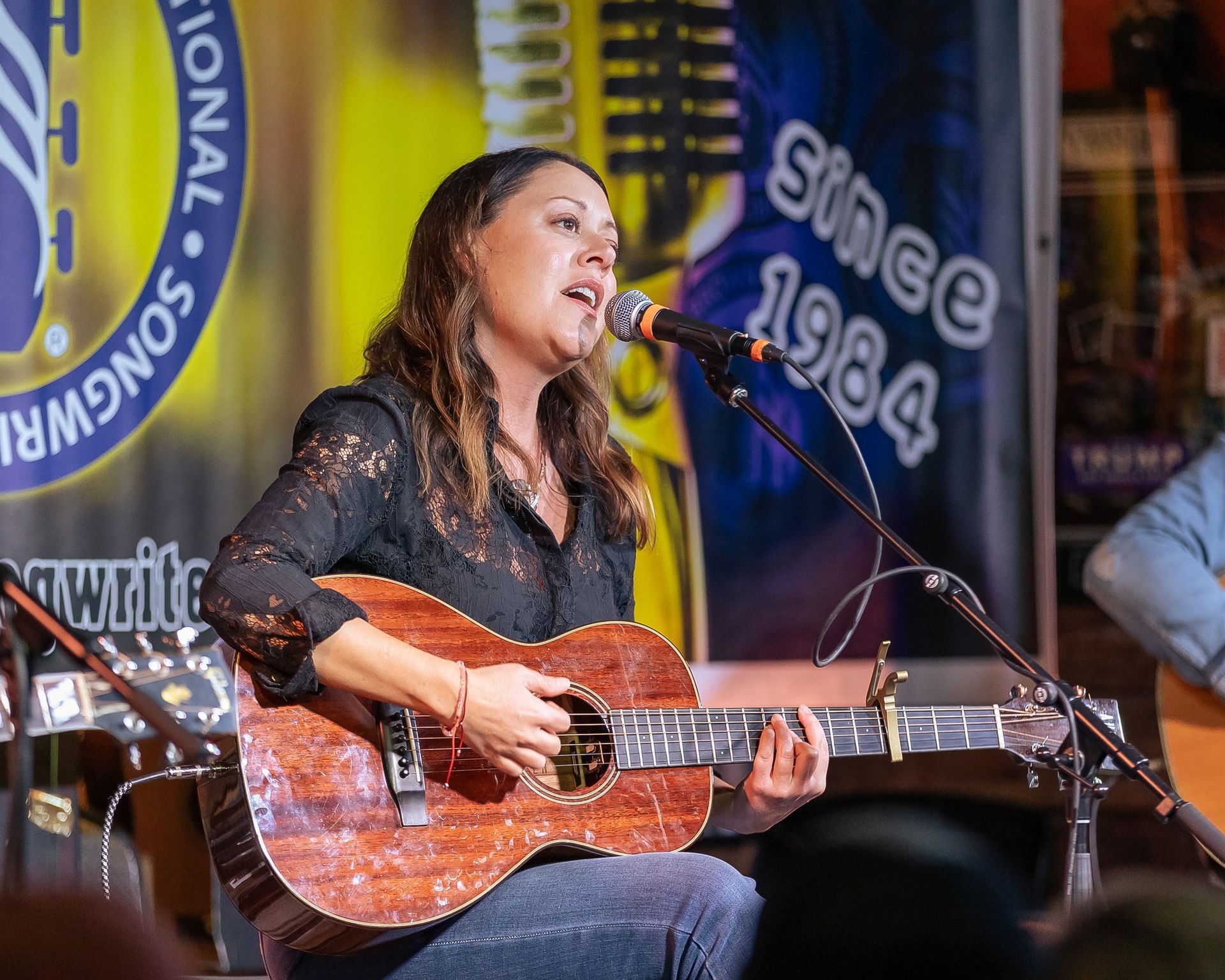 A musician singing into a microphone while playing an acoustic guitar on stage in front of a branded banner.