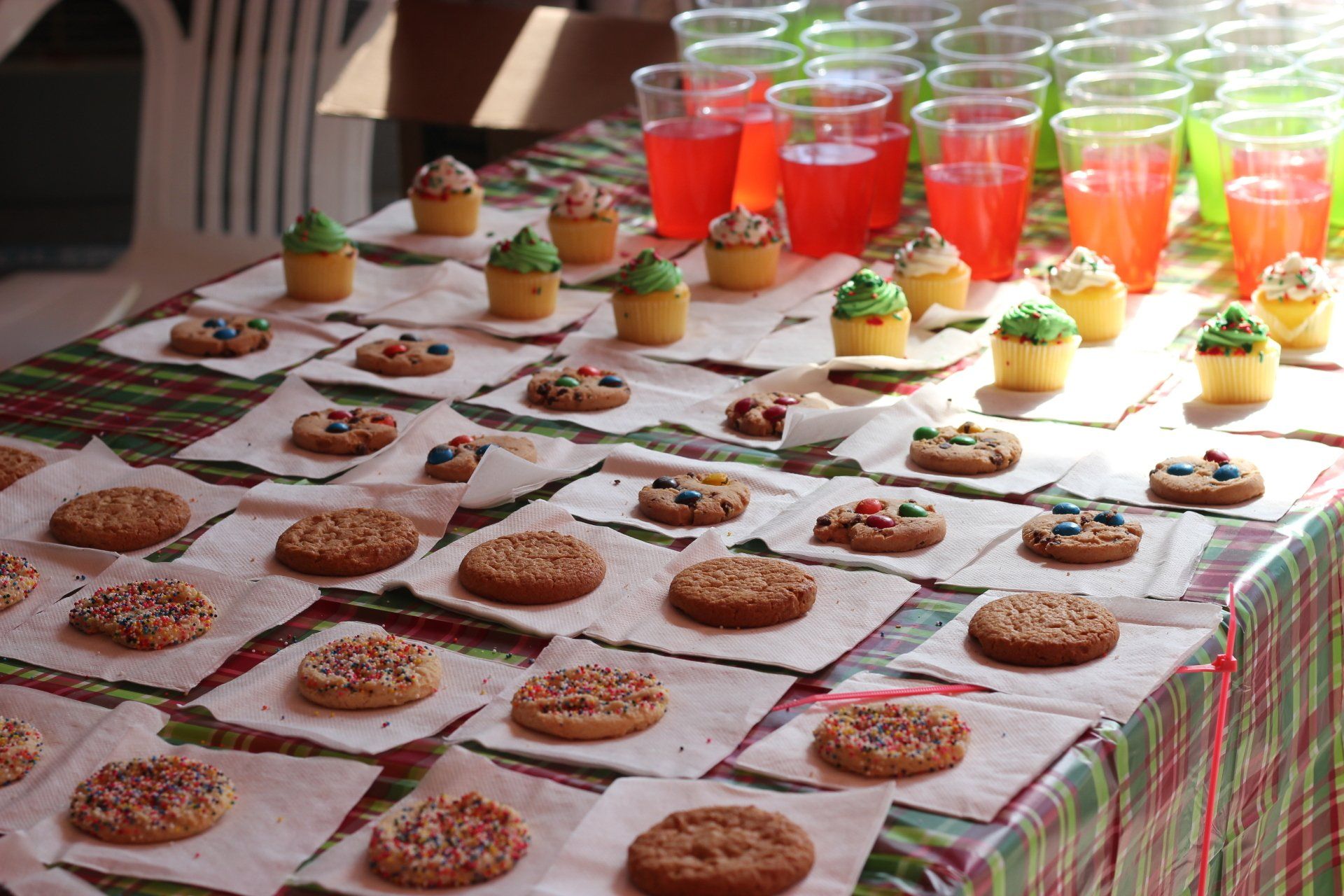 Rows of cookies on parchment paper and small frosted cupcakes arranged on a table with clear cups of red and green juice.
