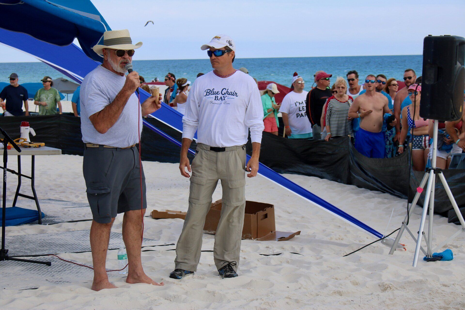 Two individuals stand under a blue canopy on a sandy beach. One speaks into a microphone, while the other watches.