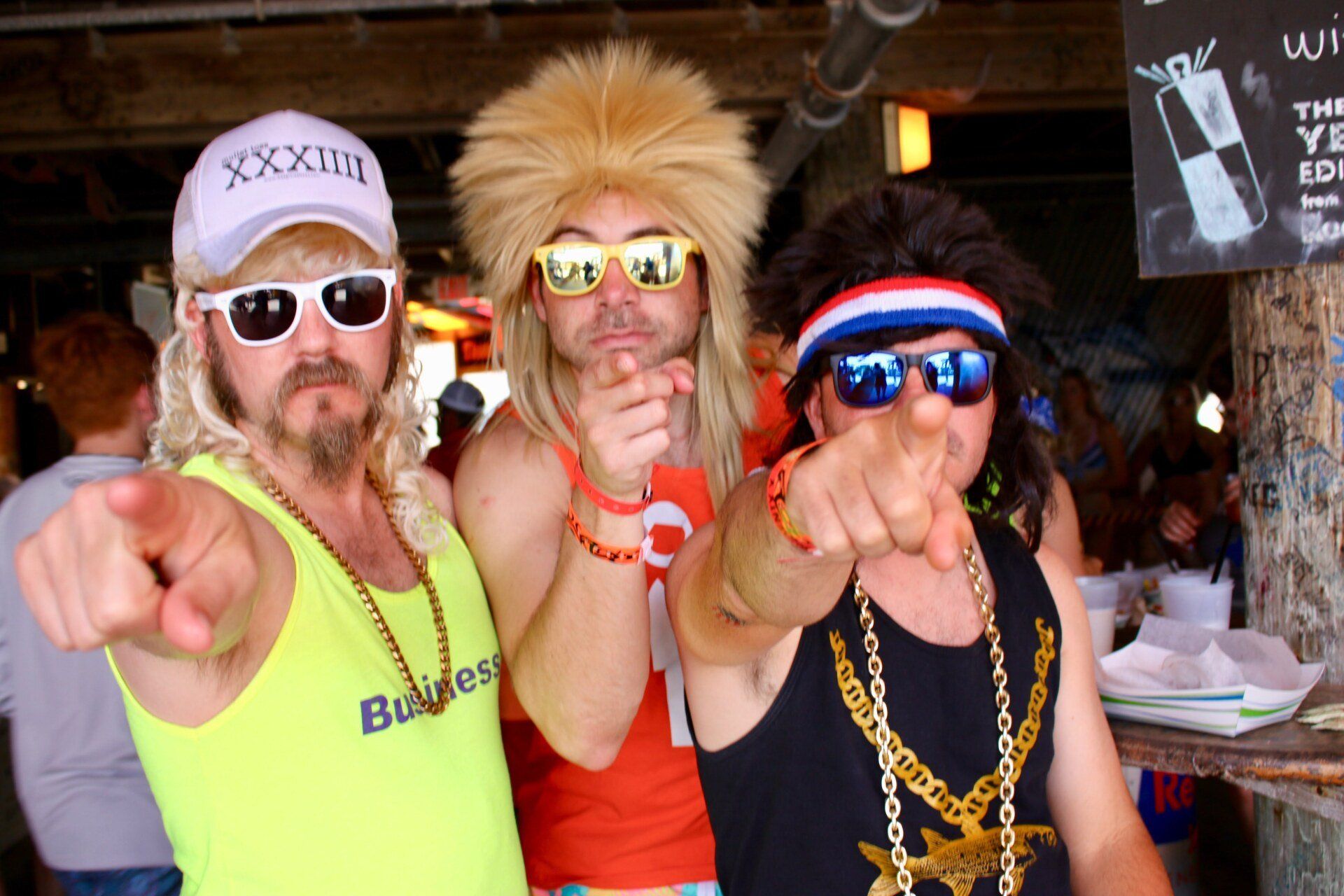 Three people in 80s-style wigs, sunglasses, and neon or graphic clothing point at the camera in a casual, outdoor setting.