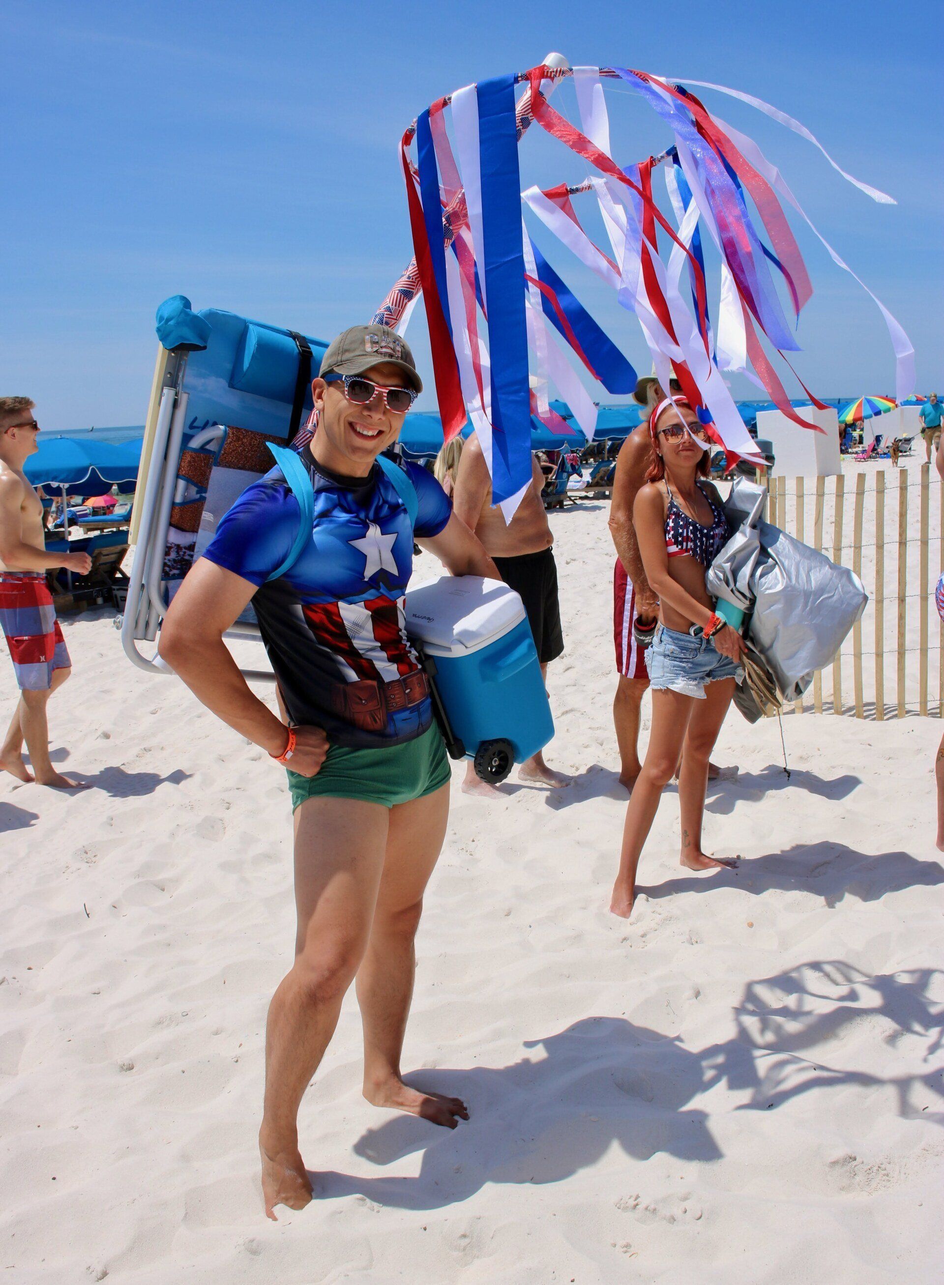 A person in a Captain America shirt and green trunks stands on a beach carrying a chair and cooler beneath festive ribbons.