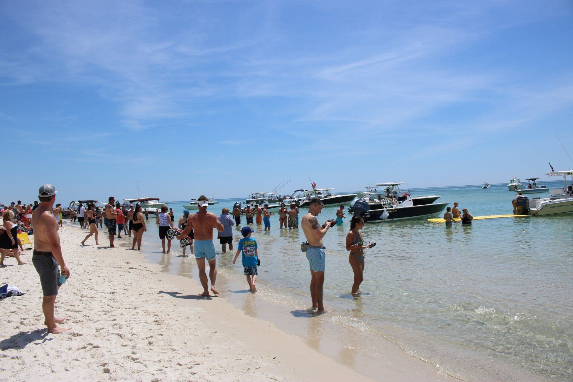 People on a sunny beach standing in the shallow water near several docked motorboats under a clear blue sky.