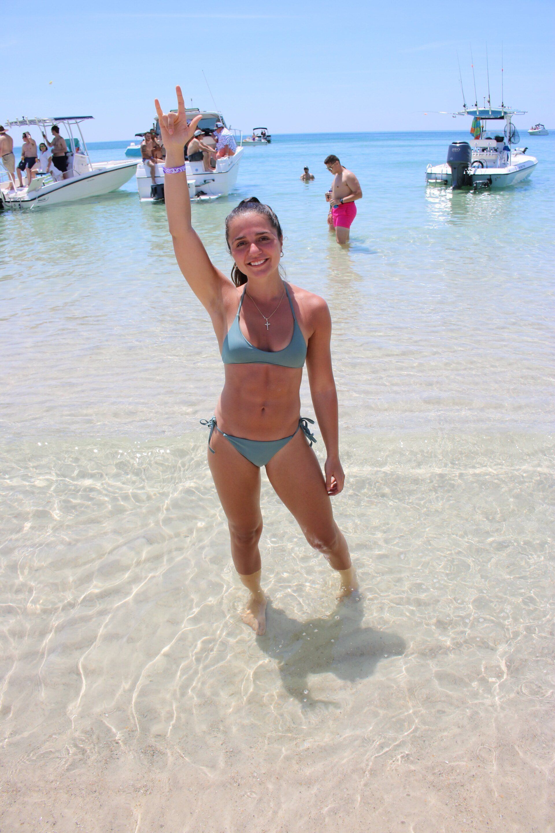 A person stands in shallow, clear ocean water, smiling and holding one hand up with fingers forming a heart gesture.