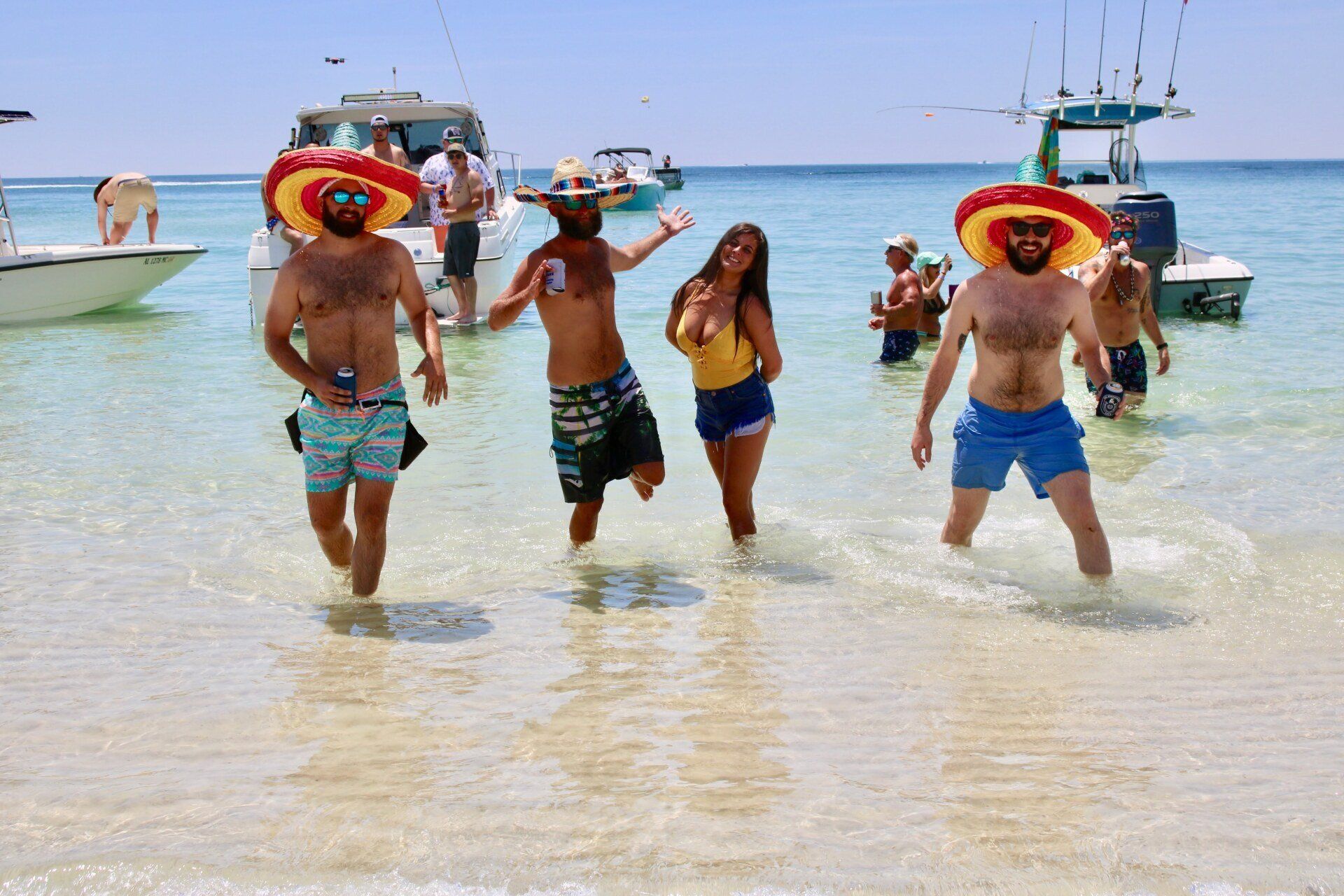 A group of people wearing hats walks through shallow ocean water near several boats on a sunny day.