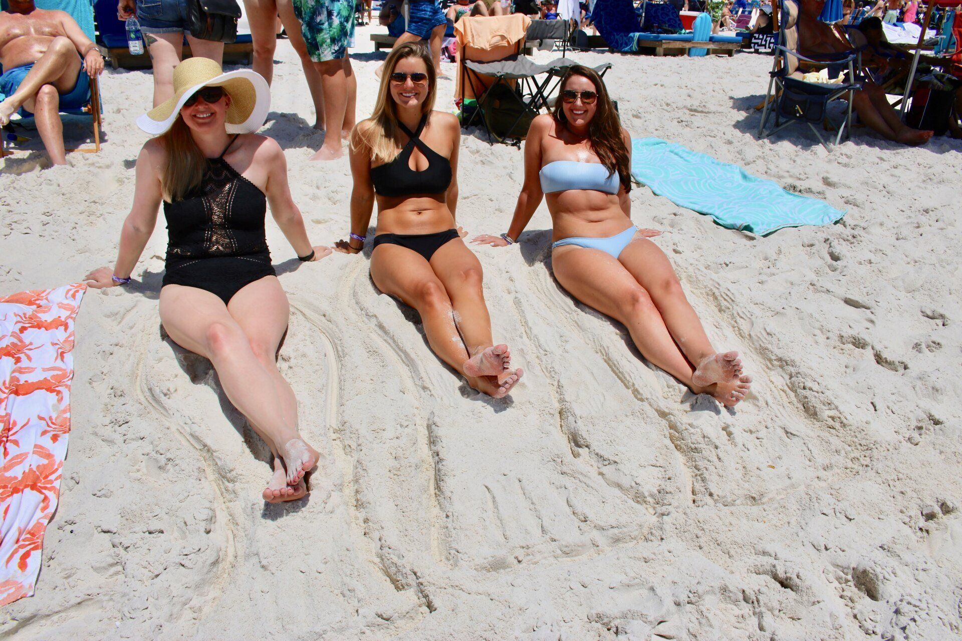 Three people sit on a sandy beach, smiling and basking in the sun while others relax in the background.