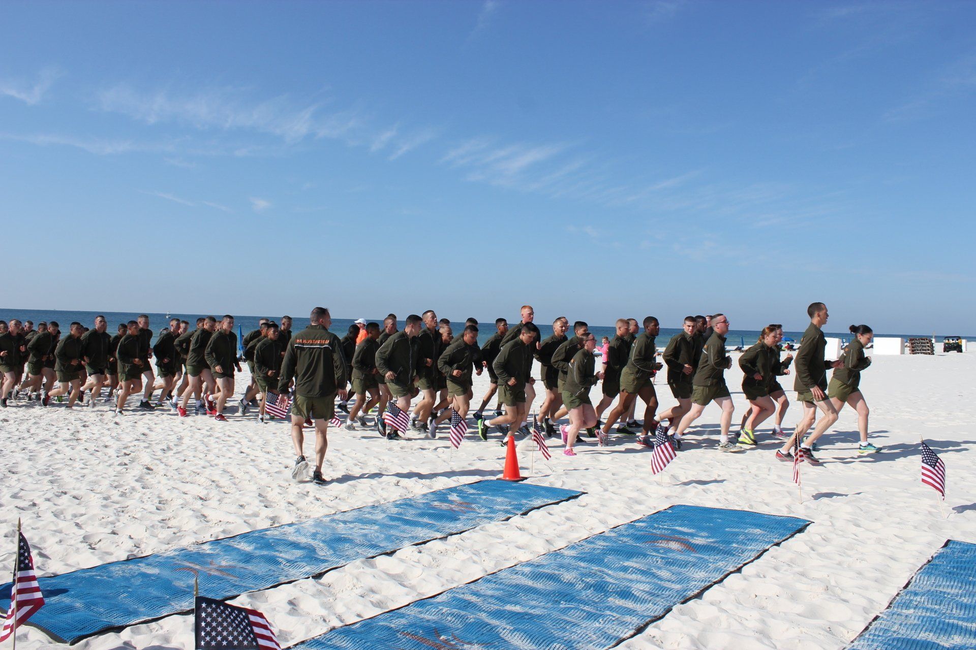 A large group of service members in matching uniforms running together on a white sand beach near the ocean.