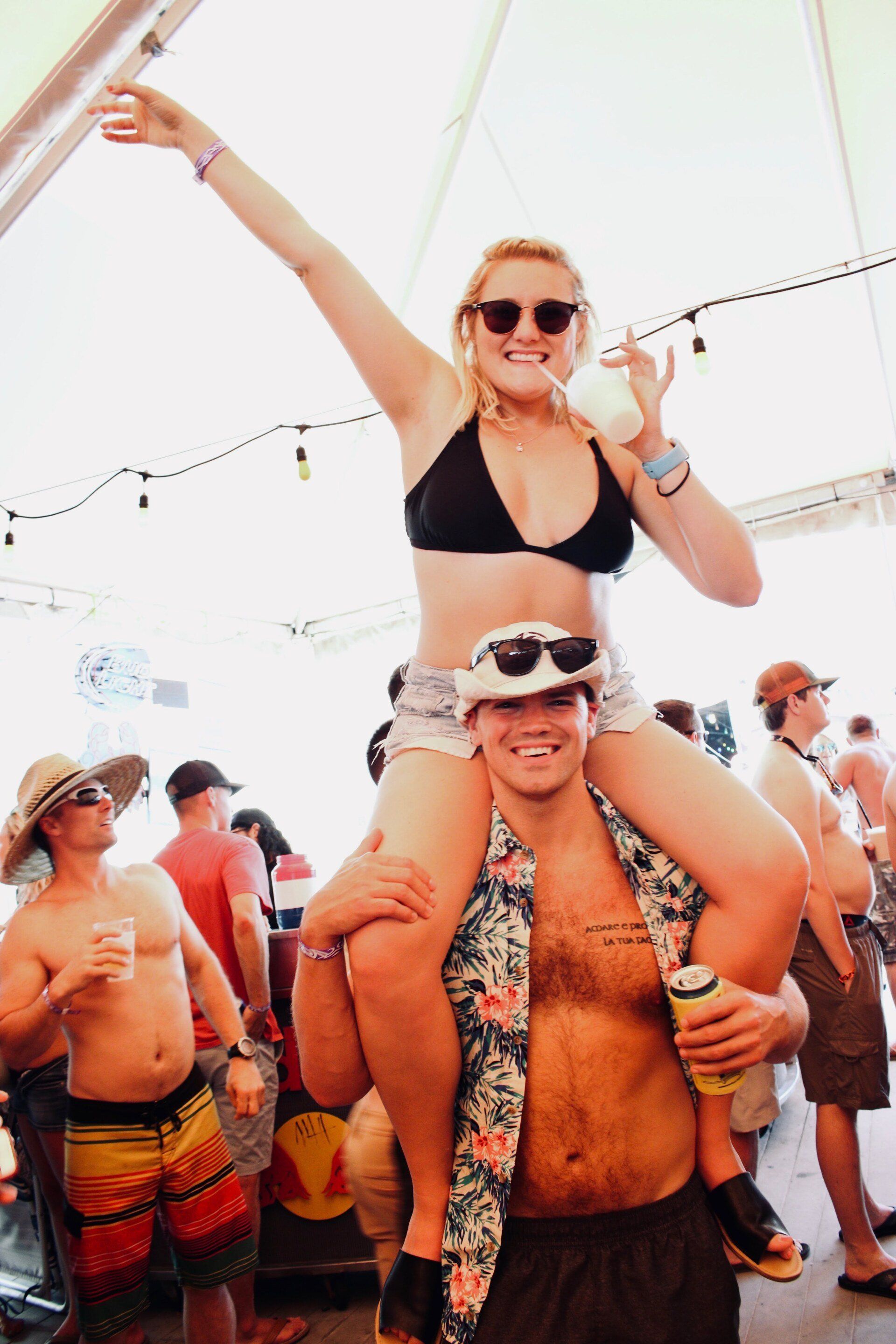 A smiling person carries another on their shoulders at a festival, with people drinking and socializing in the background.