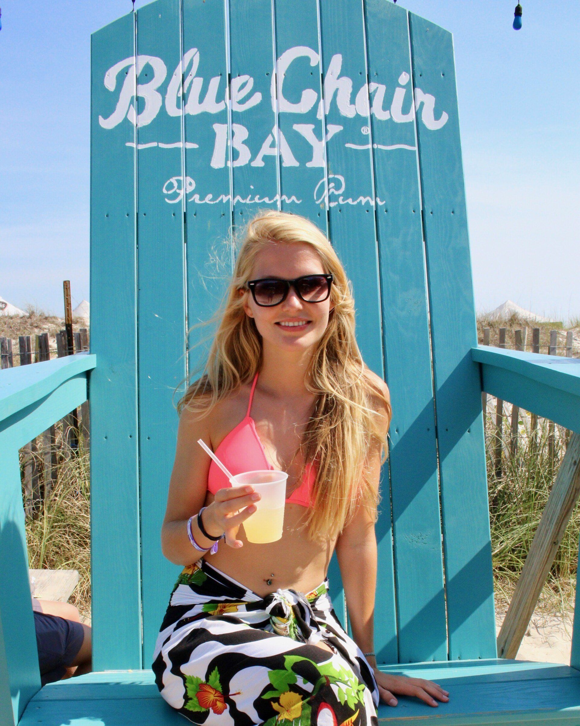 A smiling person with long blonde hair and sunglasses sits in a large, bright blue wooden chair holding a drink.
