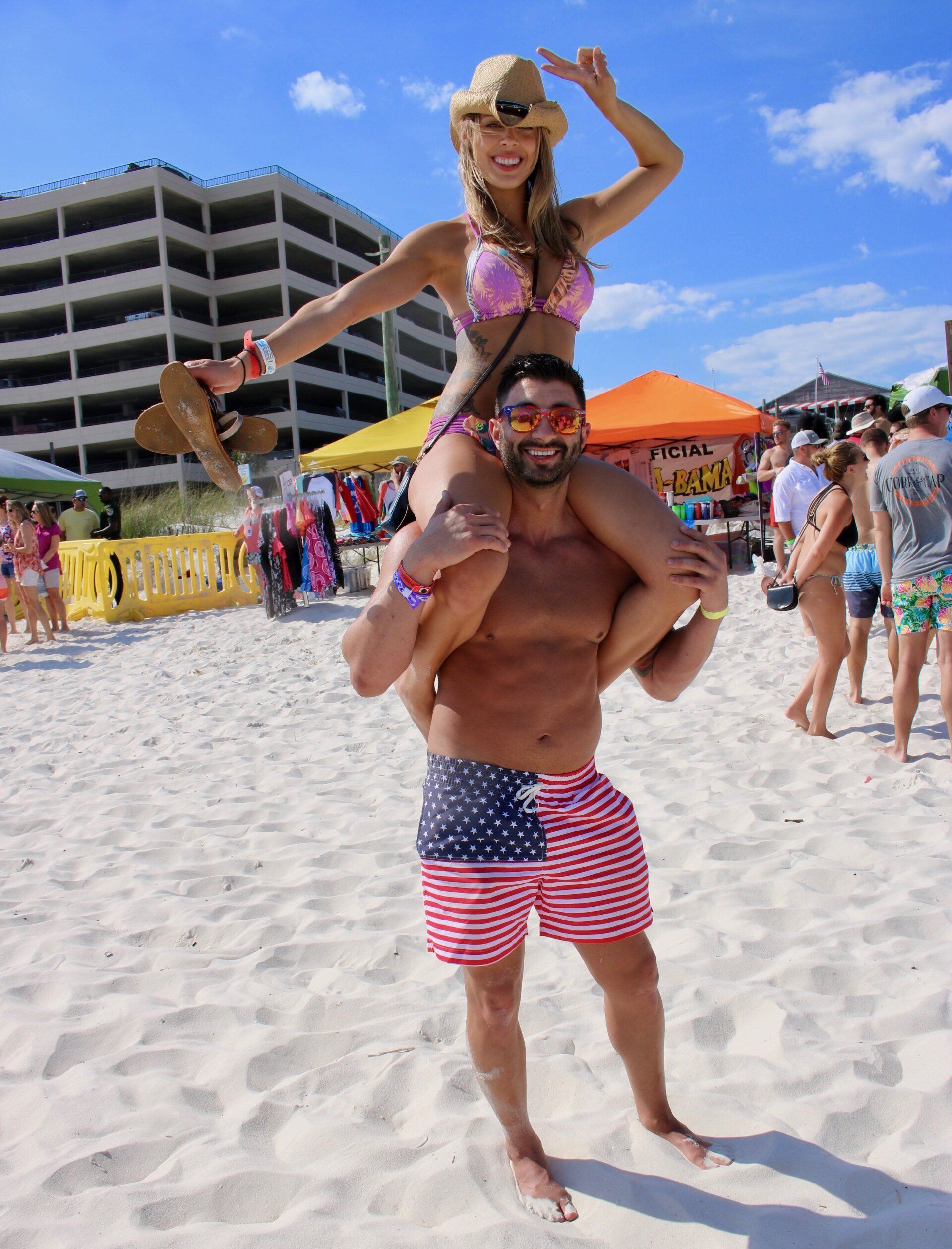 A man wearing American flag swim trunks stands on a sandy beach, carrying a woman in a bikini and cowboy hat on his shoulders.
