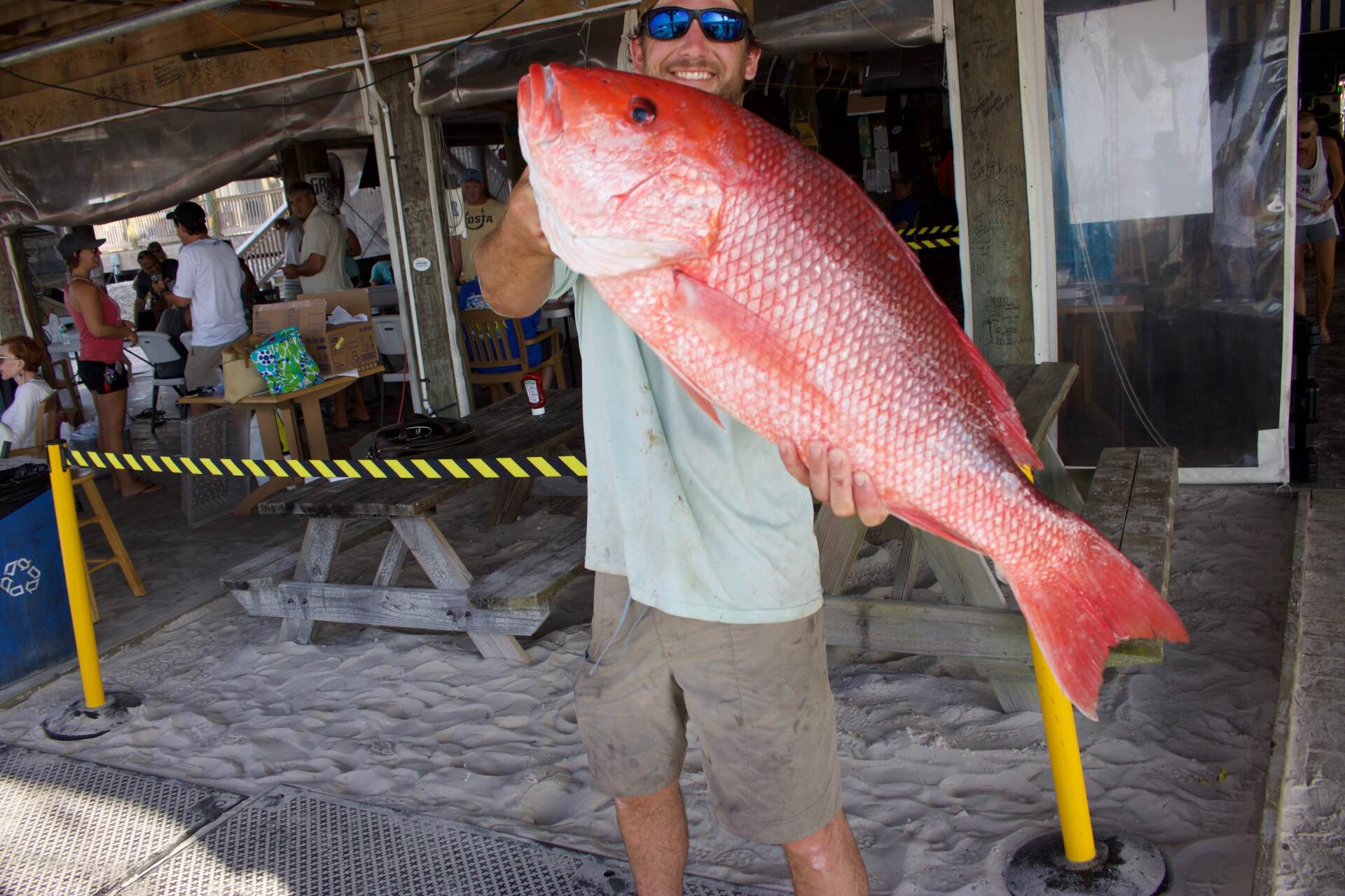 A person wearing sunglasses holds a large red snapper fish at an outdoor venue with picnic tables and sand.
