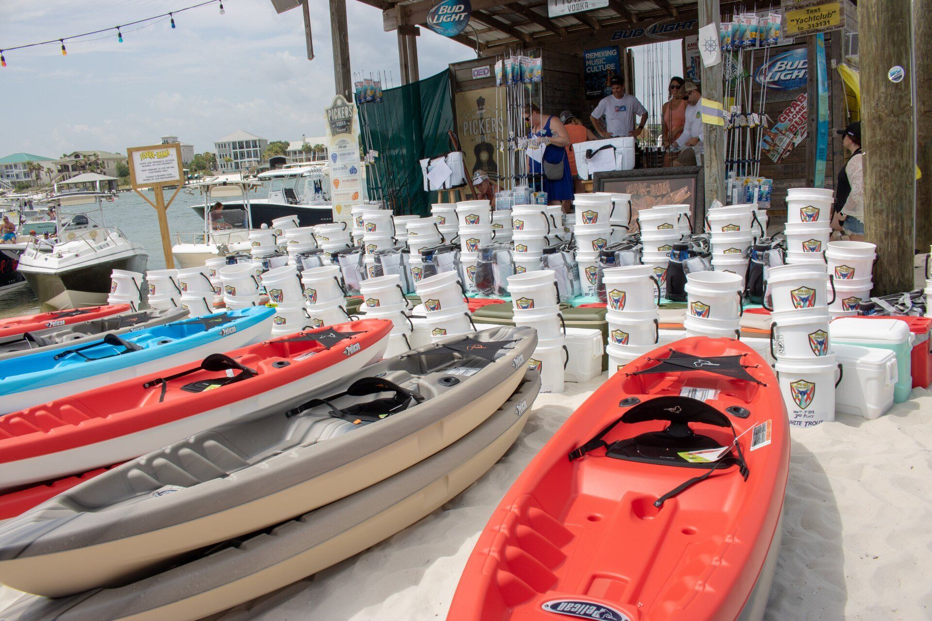 A beach rental station featuring rows of colorful kayaks on white sand, with numerous white buckets stacked in the back.