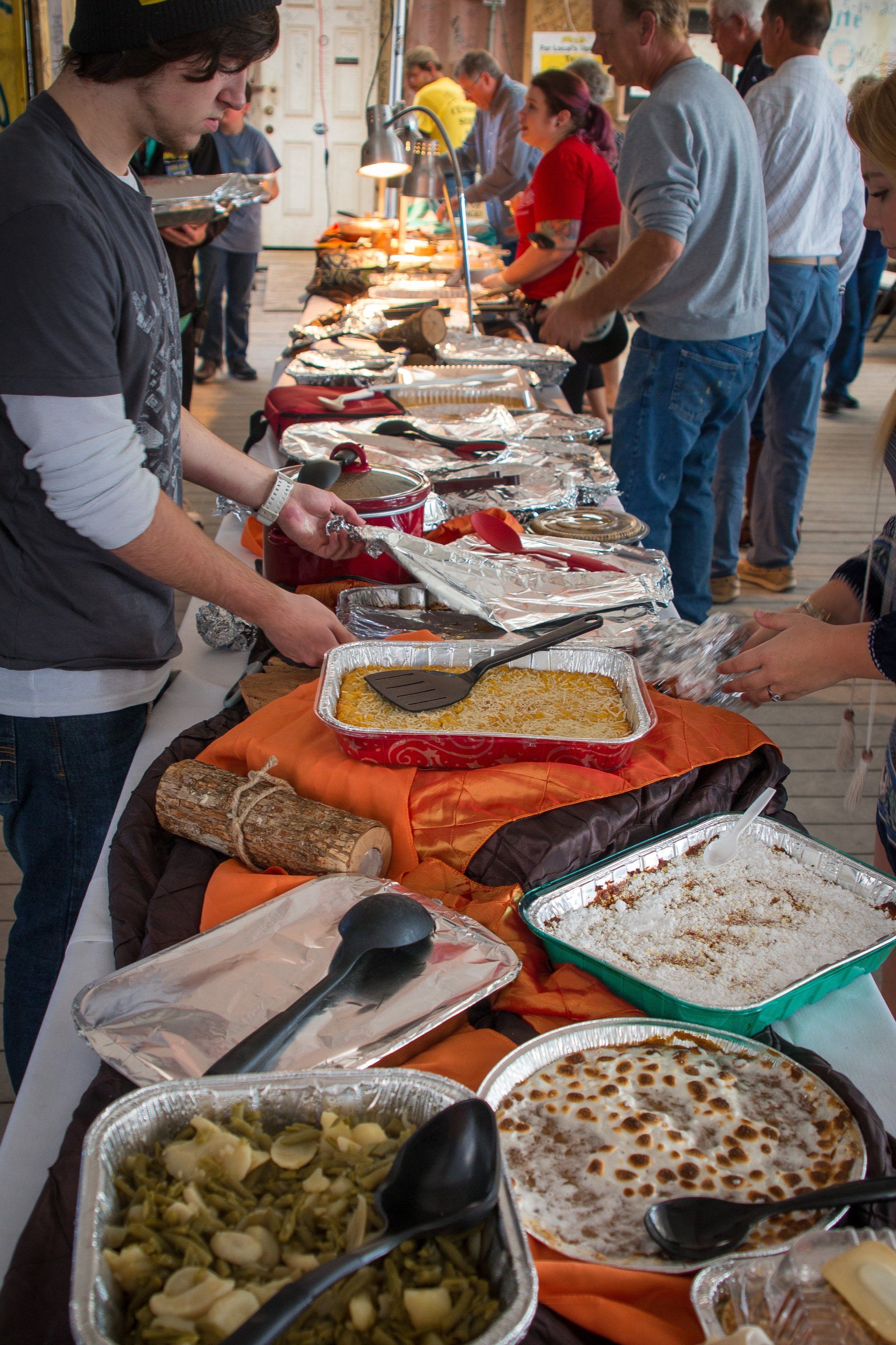 People serve themselves from a long buffet table filled with metal trays of food at an indoor gathering.