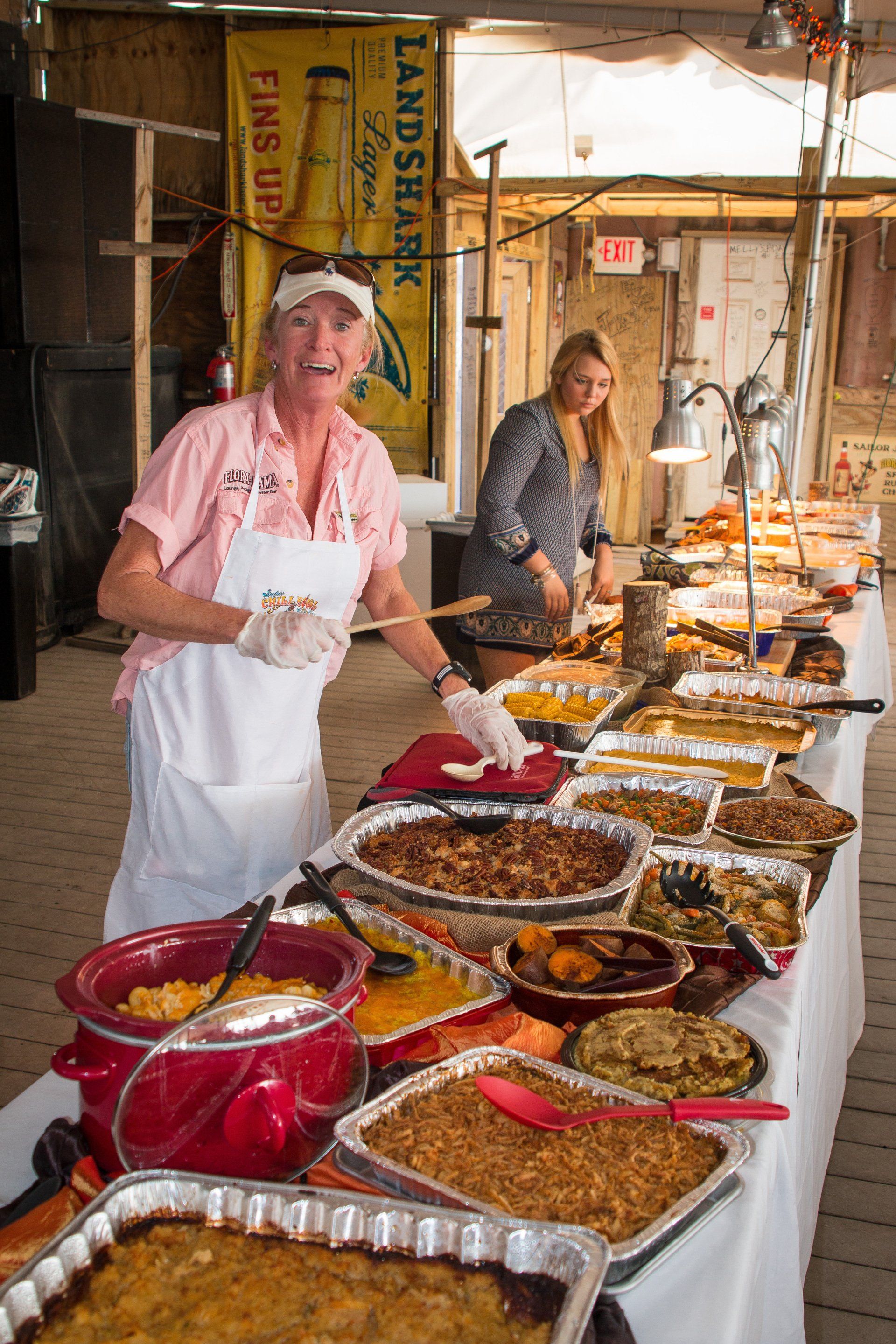 A person in a pink shirt and white apron serves food from a long buffet table under a tent with a large beer sign.