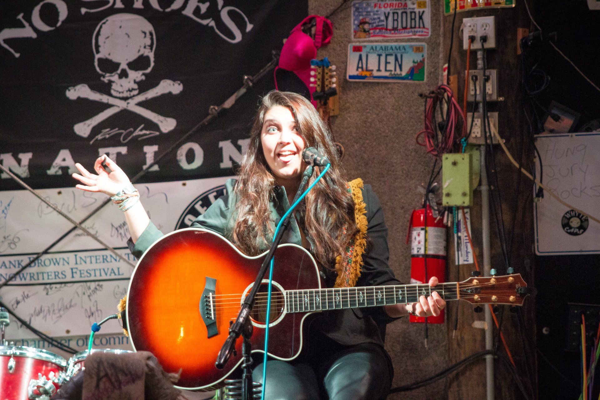 A person with long hair plays an acoustic guitar and sings into a microphone on a stage with a skull banner and decor.
