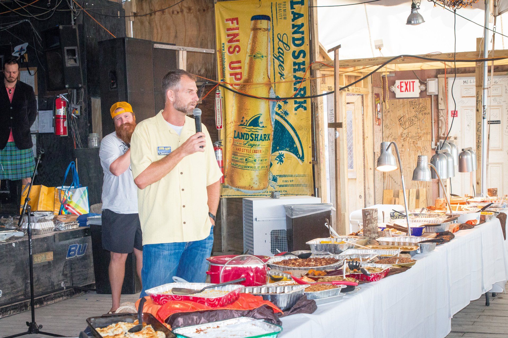 A person speaks into a microphone at an event with a buffet table, in front of a large Landshark beer banner.