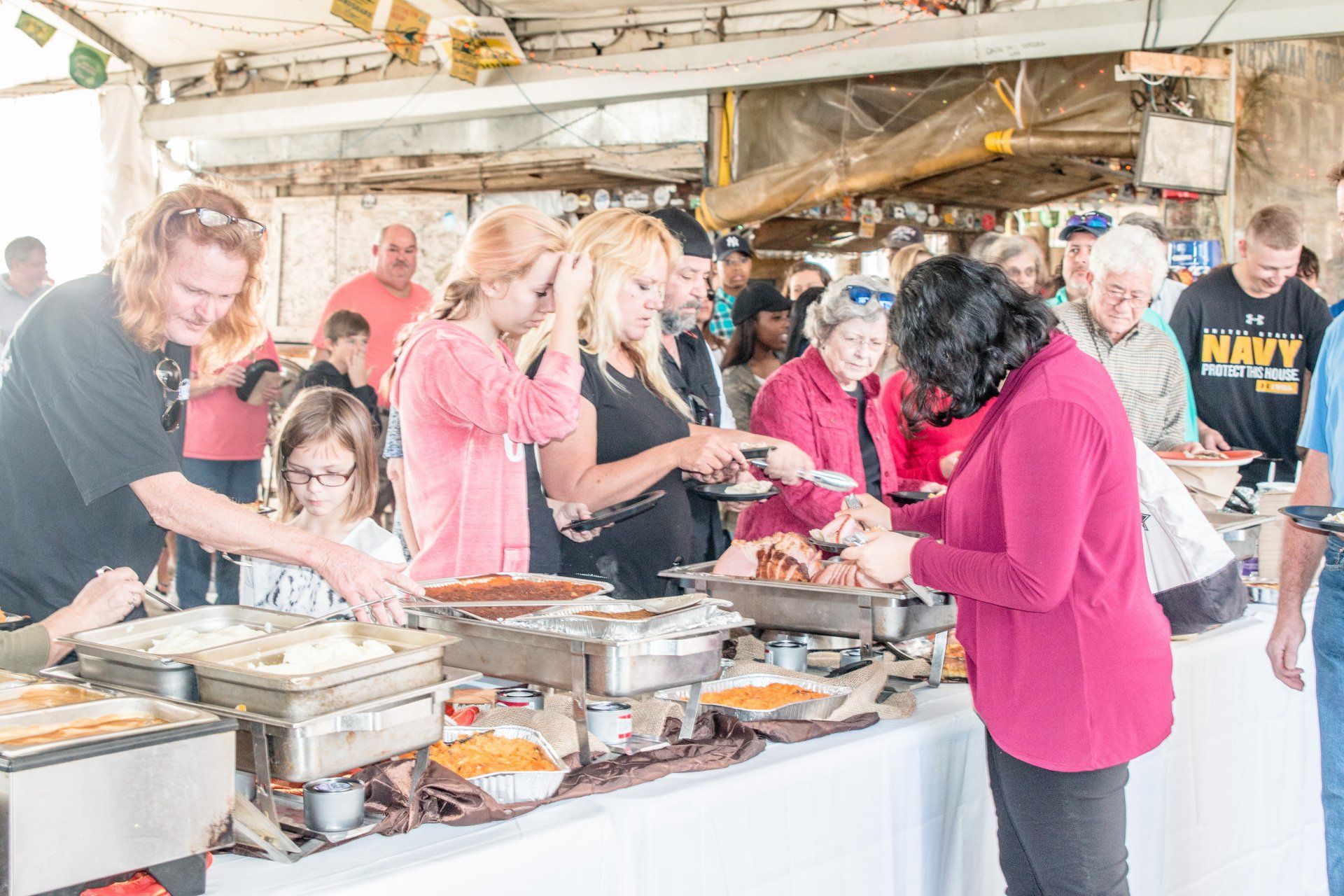 A buffet line with people in casual clothing serving themselves from metal chafing dishes under a tent.