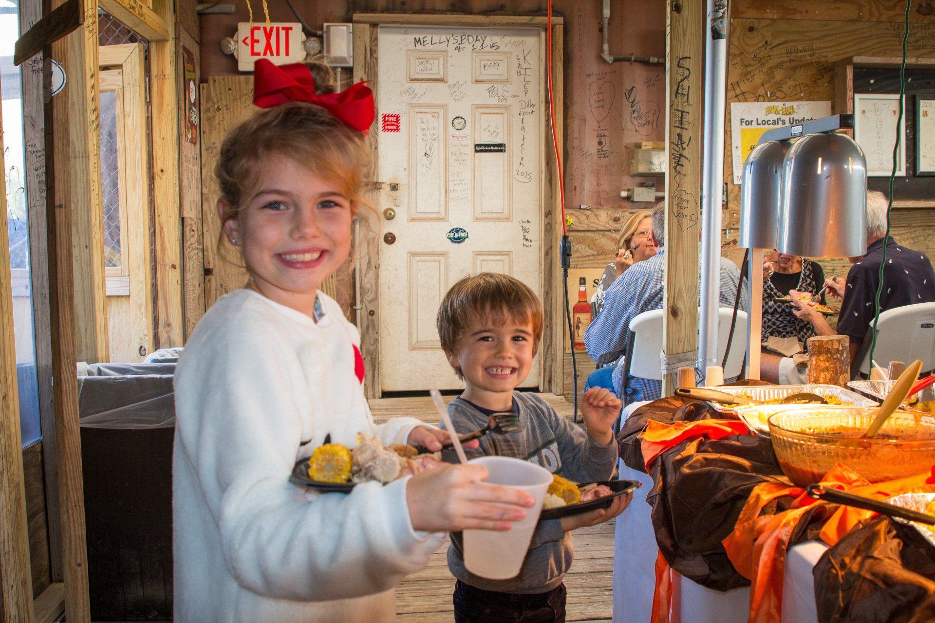 Two children holding food plates and a drink in a casual, wood-paneled room with a buffet table in the background.