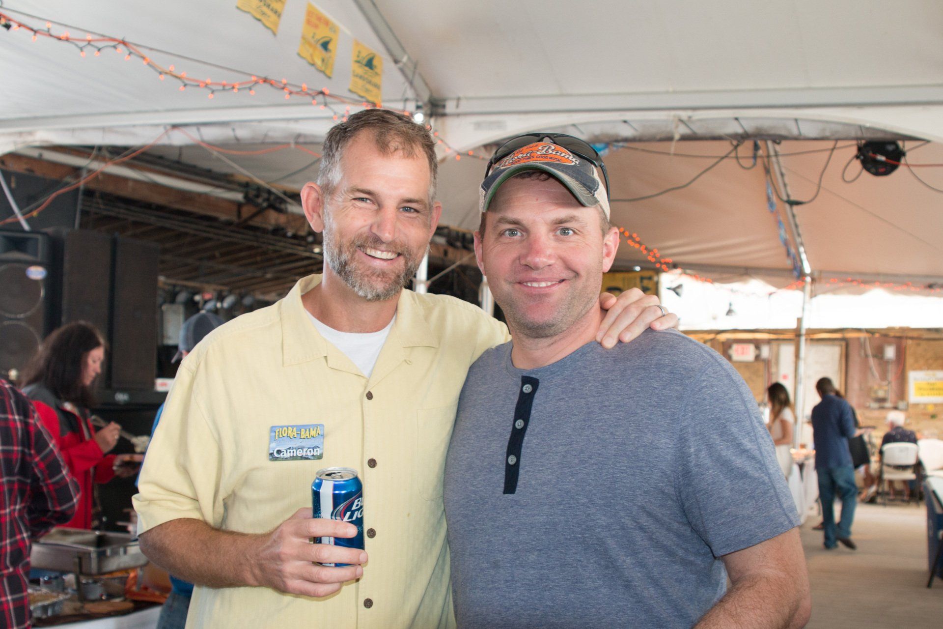Two men smile for a photo under a white tent canopy; one holds a blue beverage can.