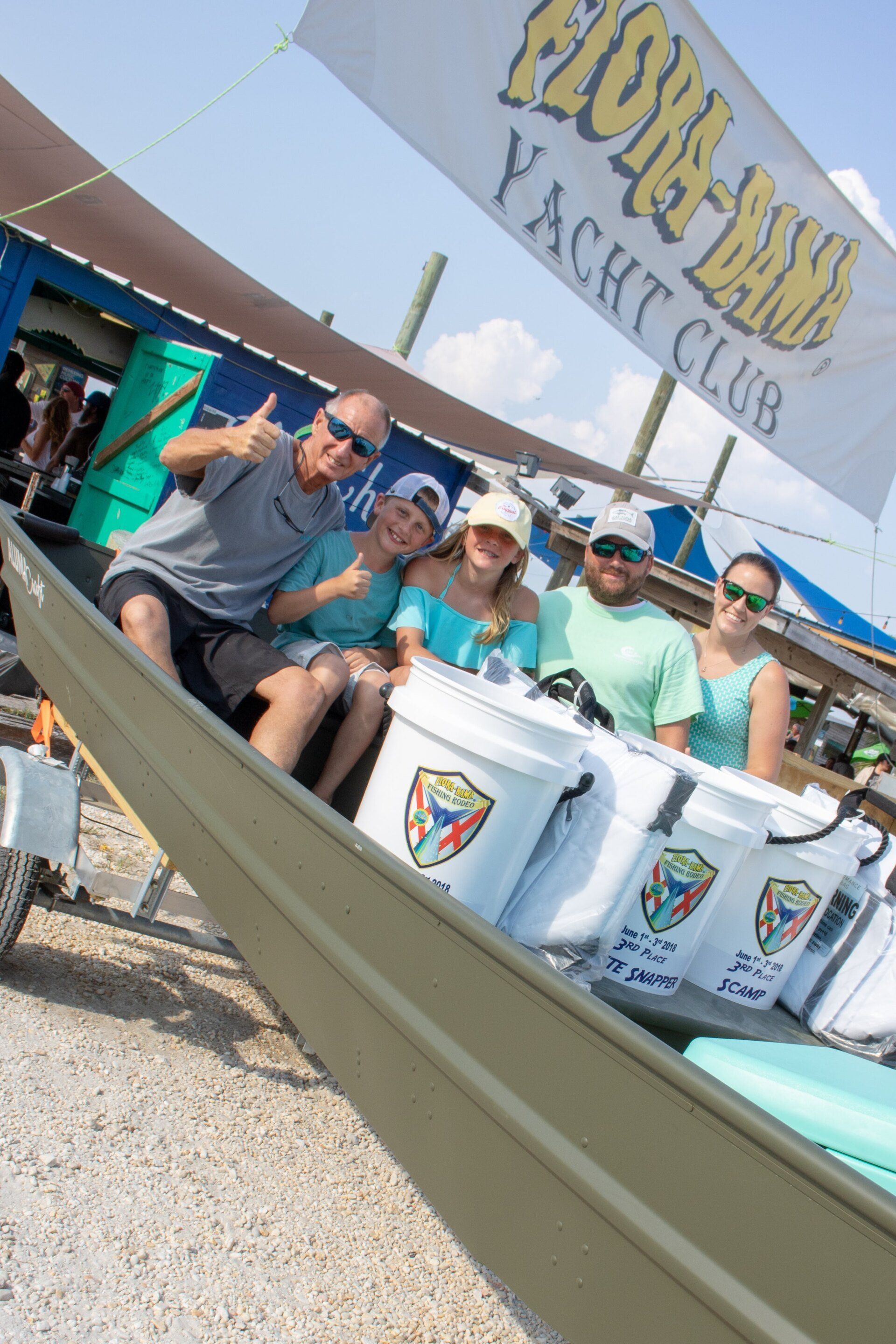 Five people pose in a small boat under a Flora-Bama Yacht Club banner on a sunny day.