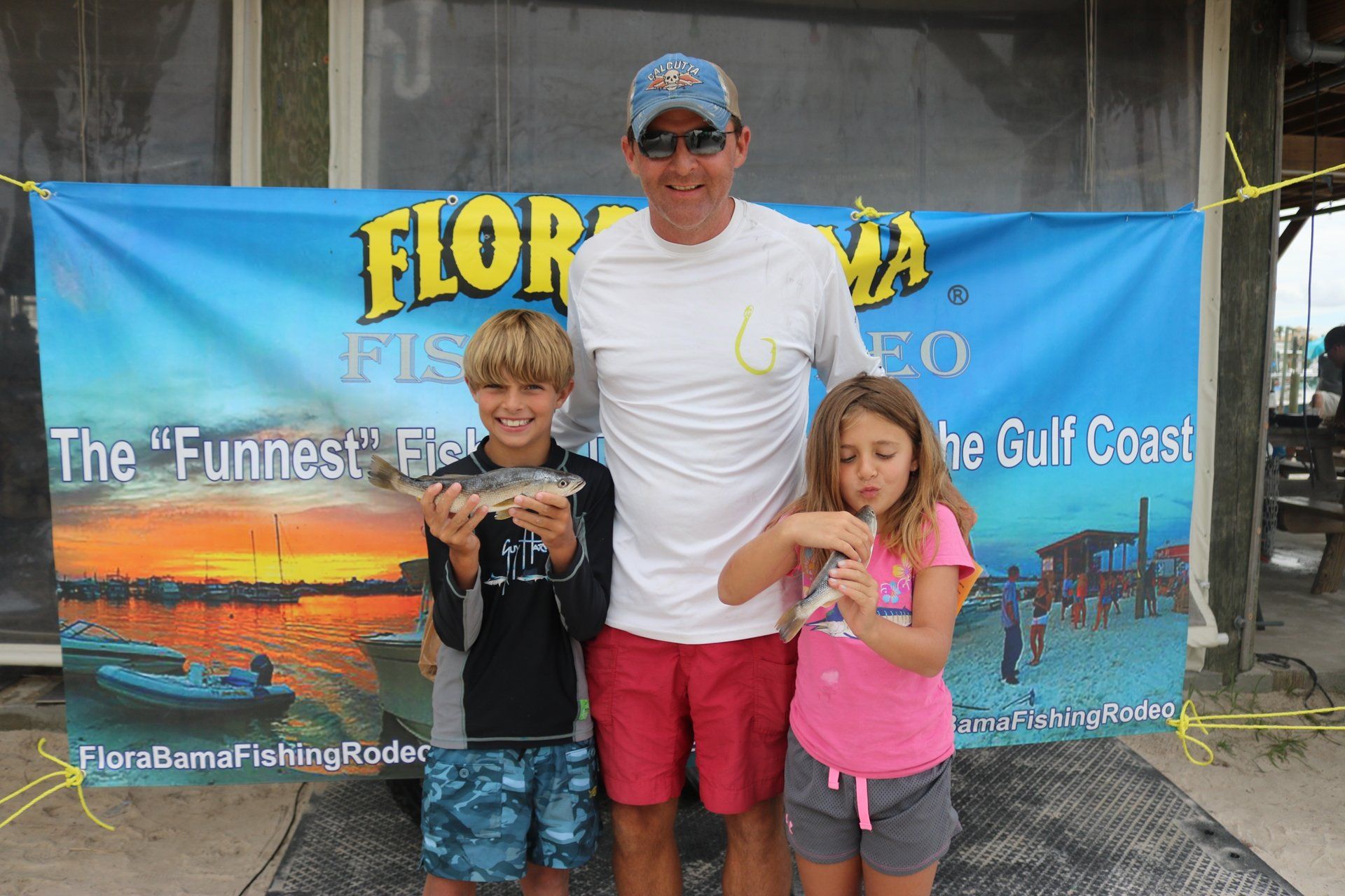 A man and two children pose holding small fish in front of a Flora-Bama Fishing Rodeo banner.